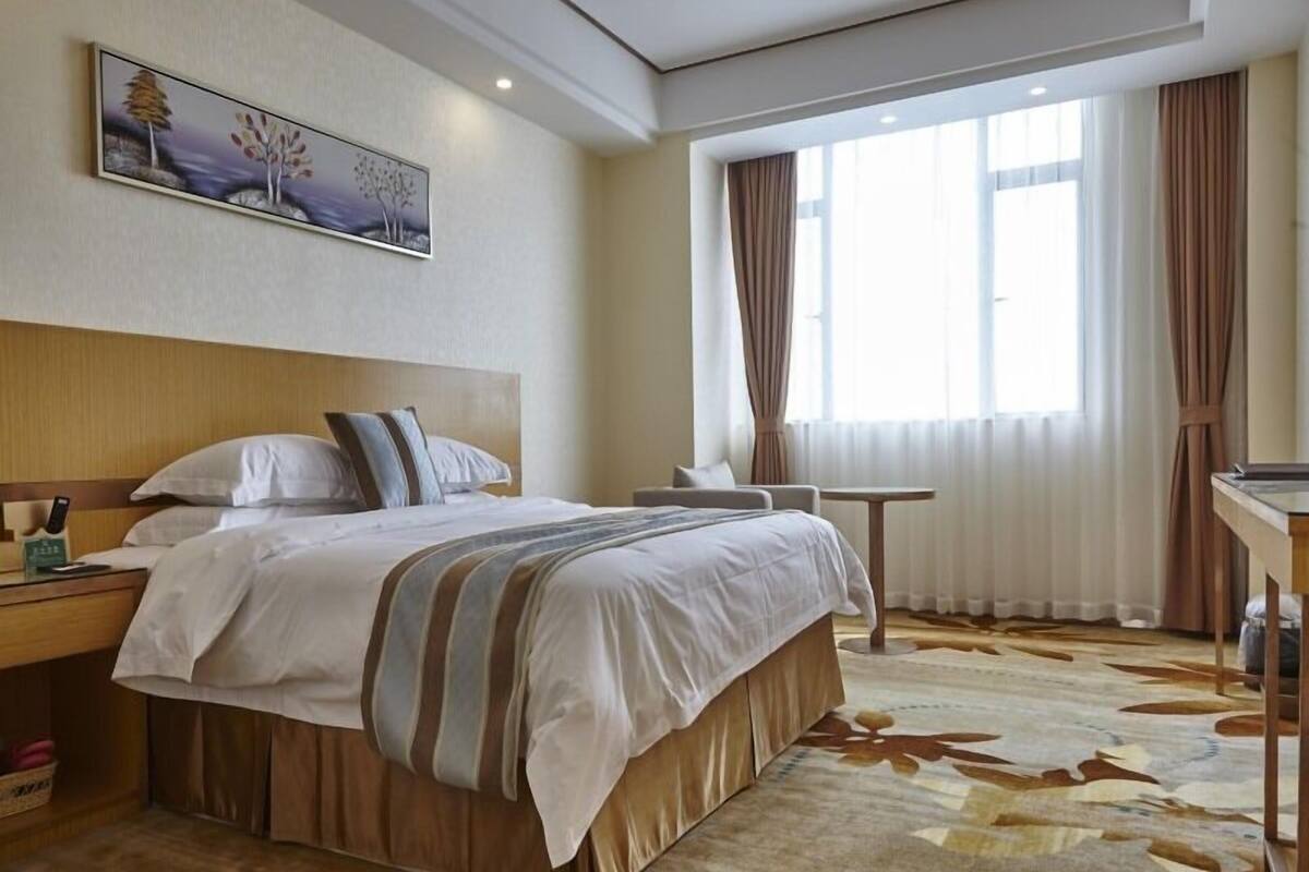 Ein Hotelzimmer mit einem großen Bett, einem Nachttisch, einem Sessel, einem kleinen Tisch und einem großen Fenster mit Vorhängen.