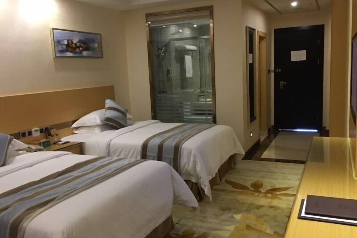 Ein Hotelzimmer mit zwei Betten, einem Nachttisch, einem Schreibtisch und einem durch eine Glastür einsehbaren Badezimmer.