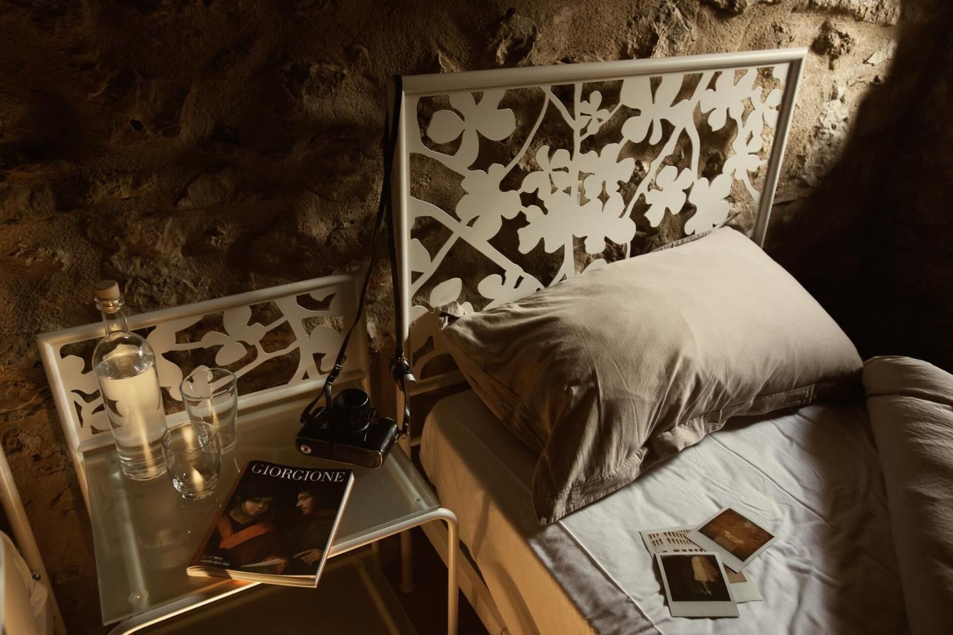 A bed with a white headboard and a pillow, a bedside table with a book titled 'Giorgione', a bottle, and glasses.