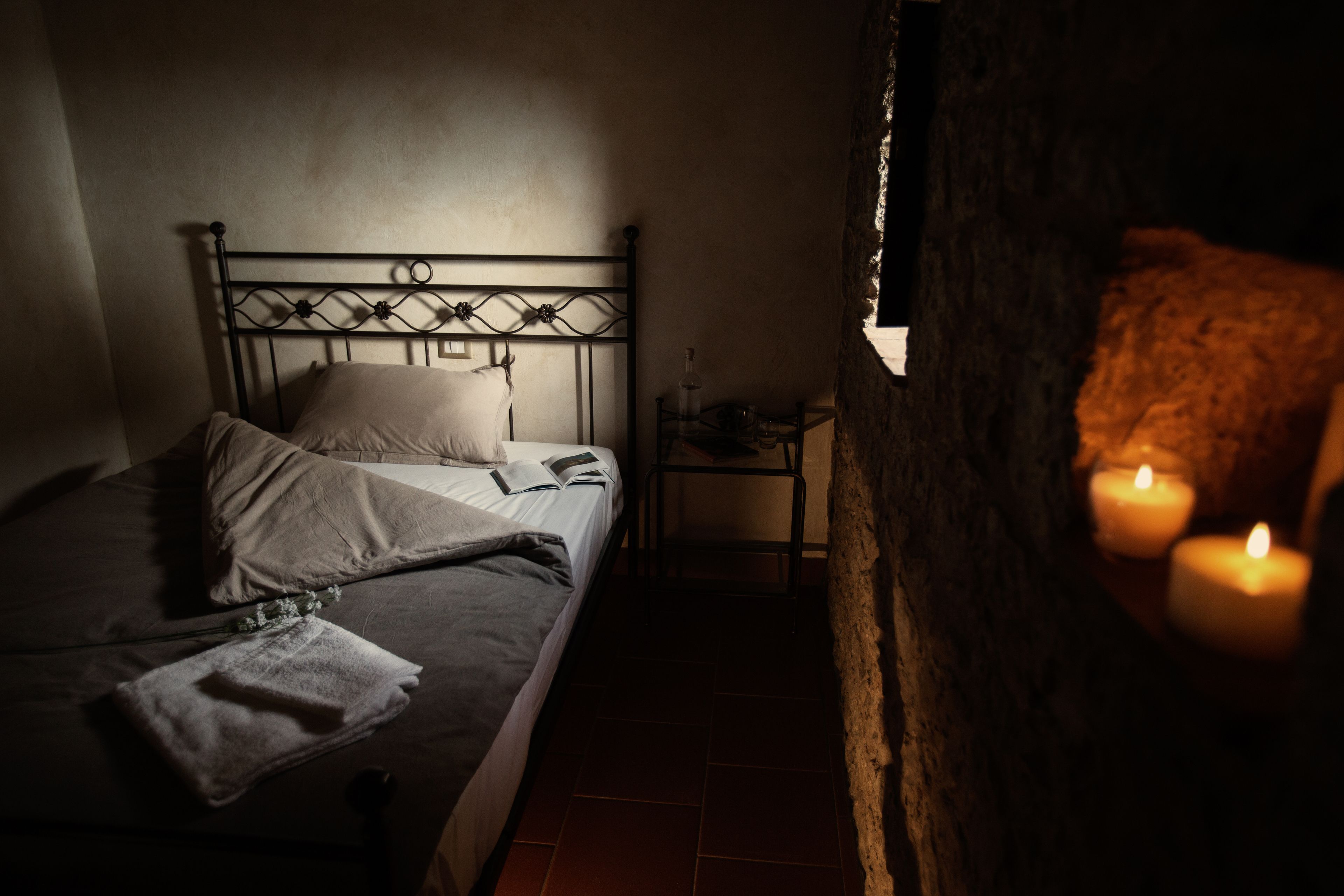 Un dormitorio con poca luz, estructura de cama metálica, mesita de noche con lámpara y dos velas encendidas en una mesita.