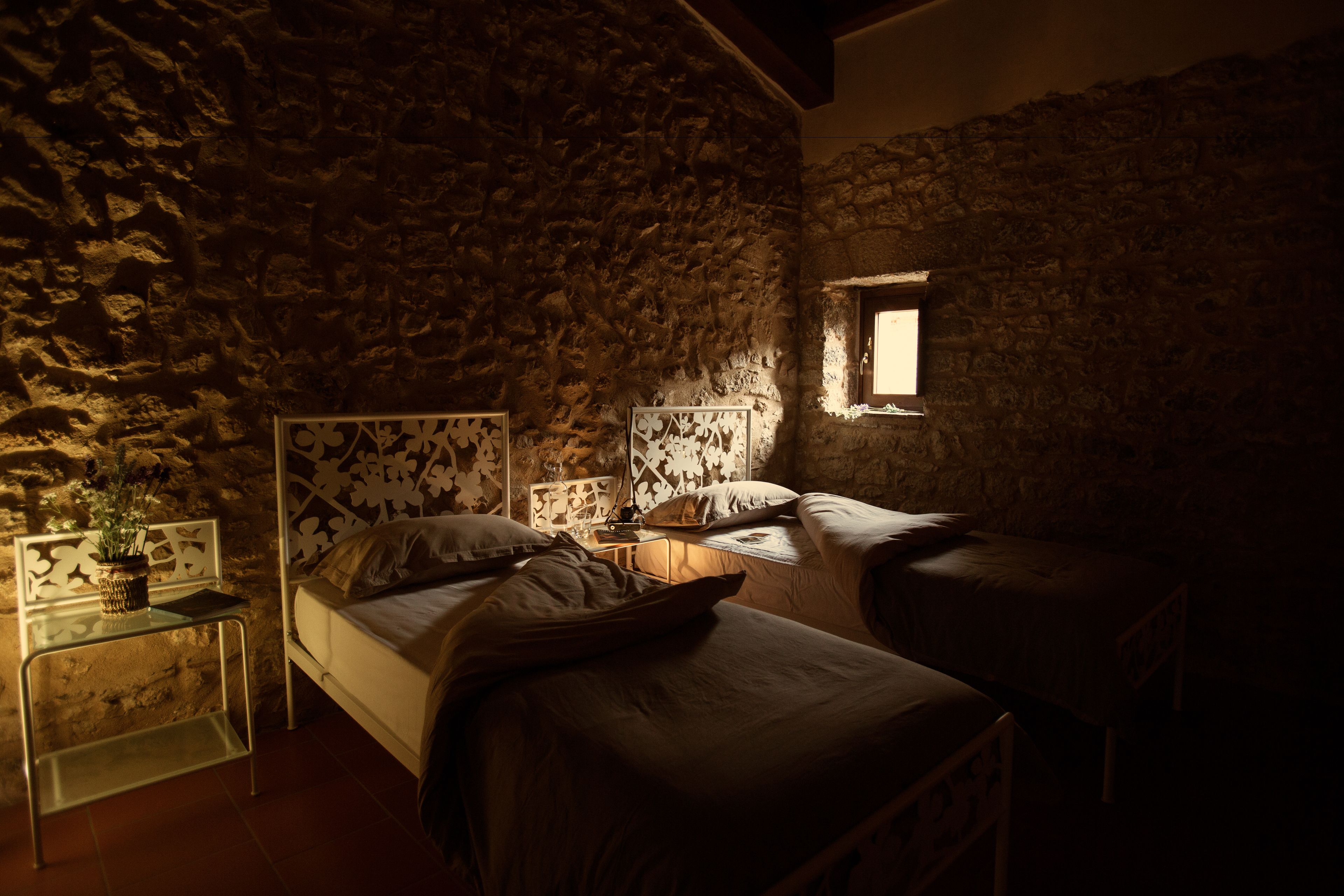 Een zwak verlichte kamer met een stenen muur, een bed met hoofdeinde, een nachtkastje met lamp en een klein raam.