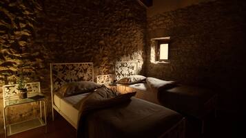 A dimly lit room with a stone wall, a bed with a headboard, a bedside table with a lamp, and a small window.