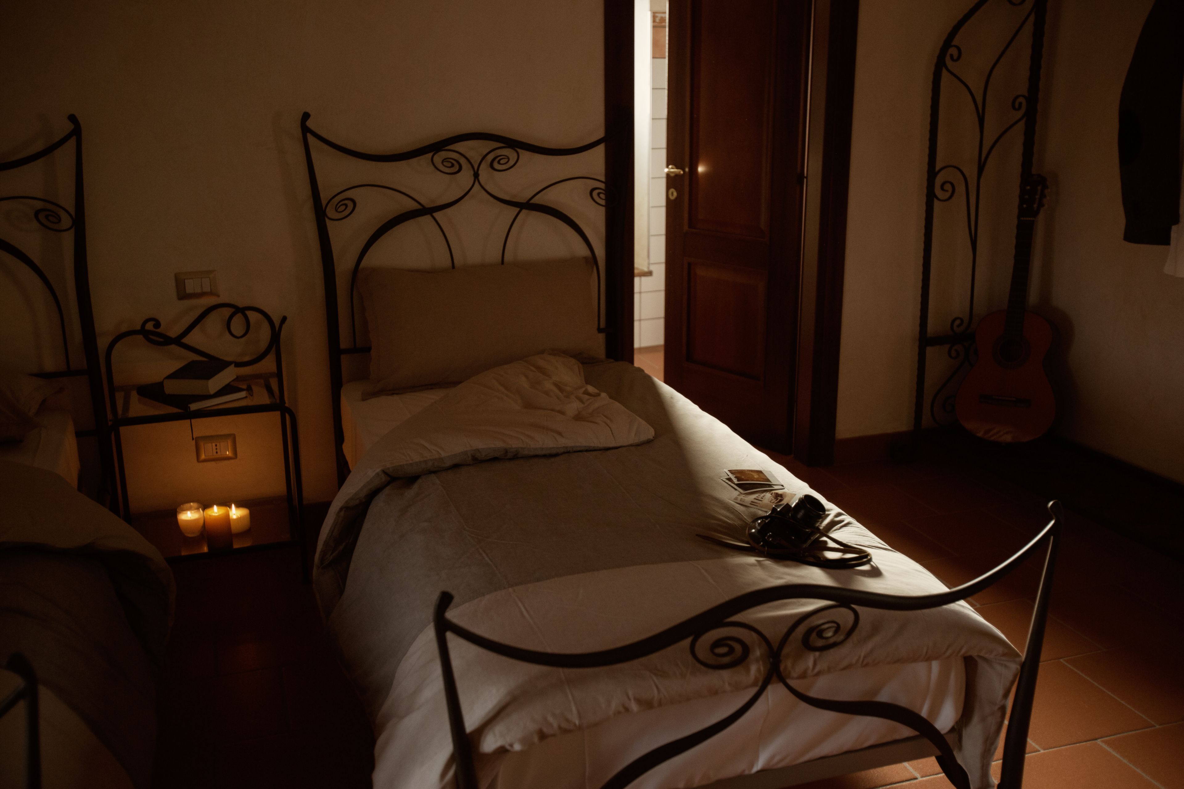 A dimly lit bedroom with a single bed, a nightstand with candles, a guitar, and a door.