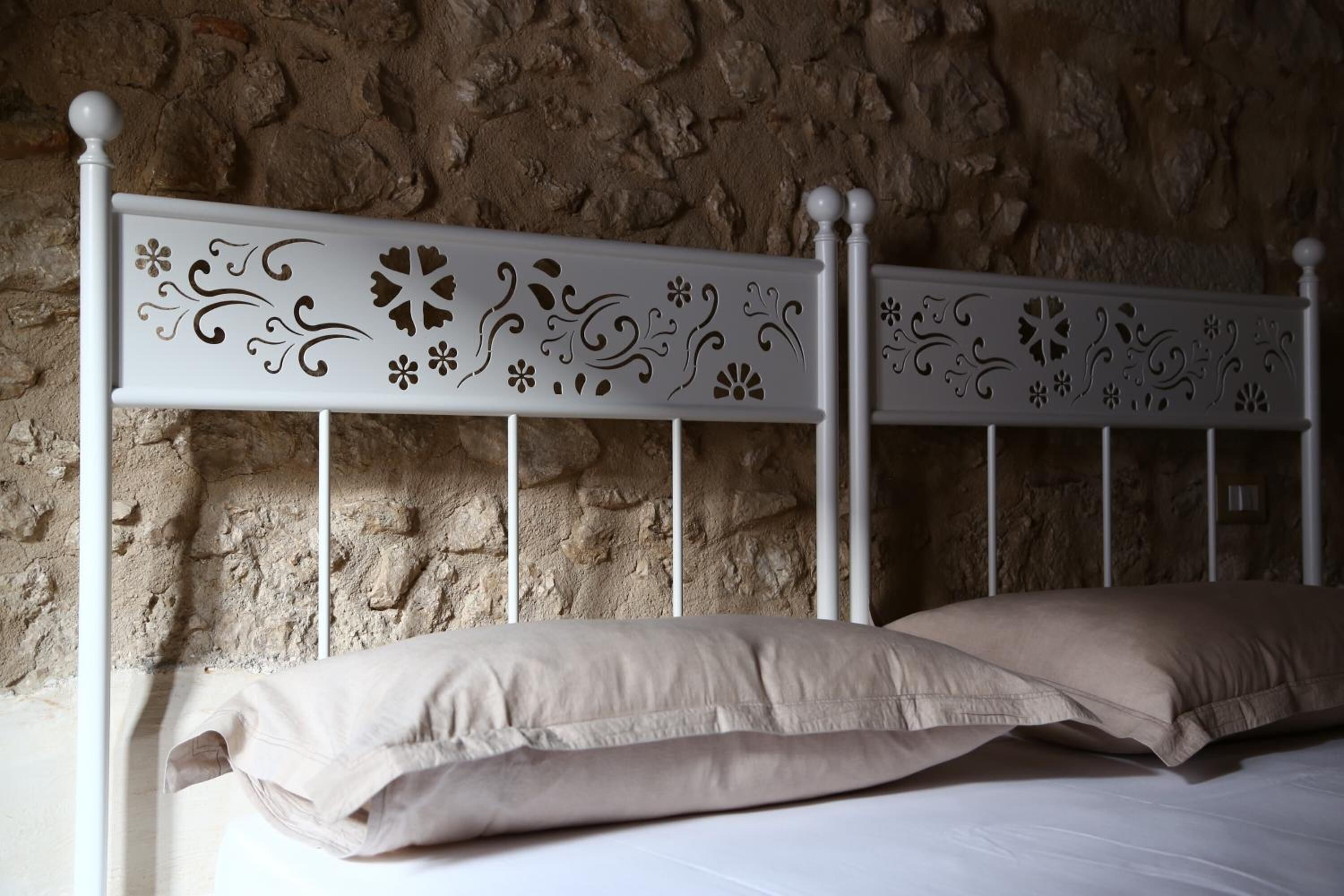 A bed with a white metal headboard featuring decorative cut-out patterns, set against a textured stone wall.