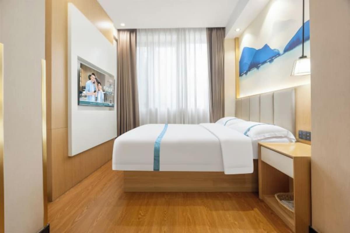 A hotel room with a bed, a bedside table, a mirror, a window with curtains, and a wall-mounted TV.