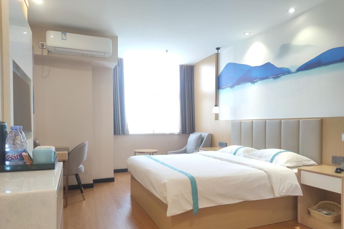 A hotel room with a bed, a desk, a chair, an air conditioning unit, and a window with curtains.