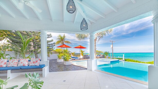 A poolside area with a view of the ocean, a seating area with cushions, and two red umbrellas. - Villa North Winds Orange Hill (Nassau)