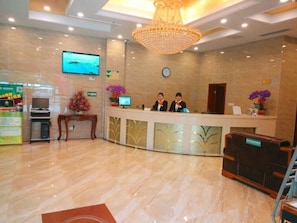 Un'area della reception di un hotel con pavimento in marmo, un lampadario, un banco reception, una sveglia e un televisore appeso al muro.