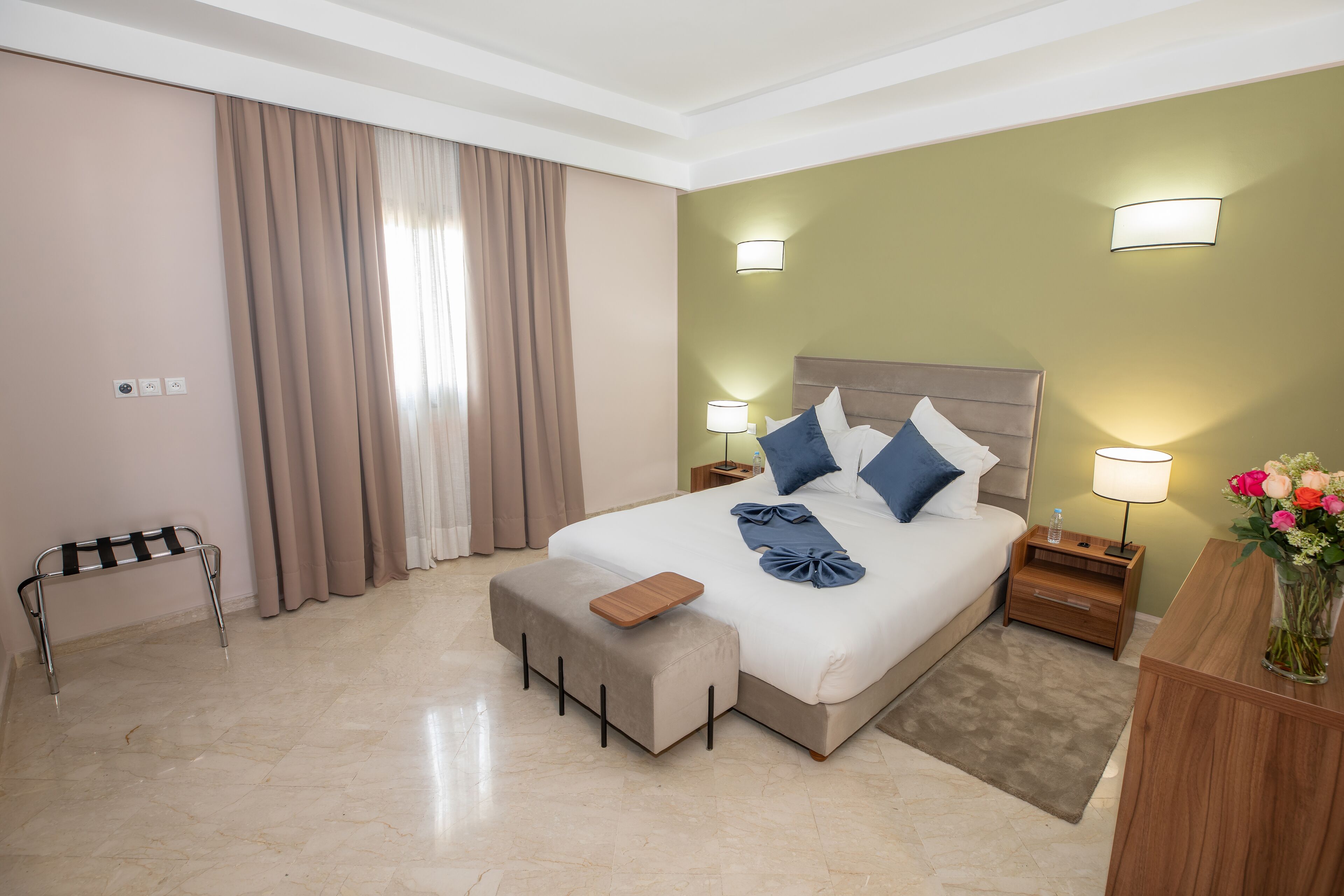 Habitació d'hotel amb un llit gran, una banca, una tauleta de nit i un escriptori amb un ram de flors.