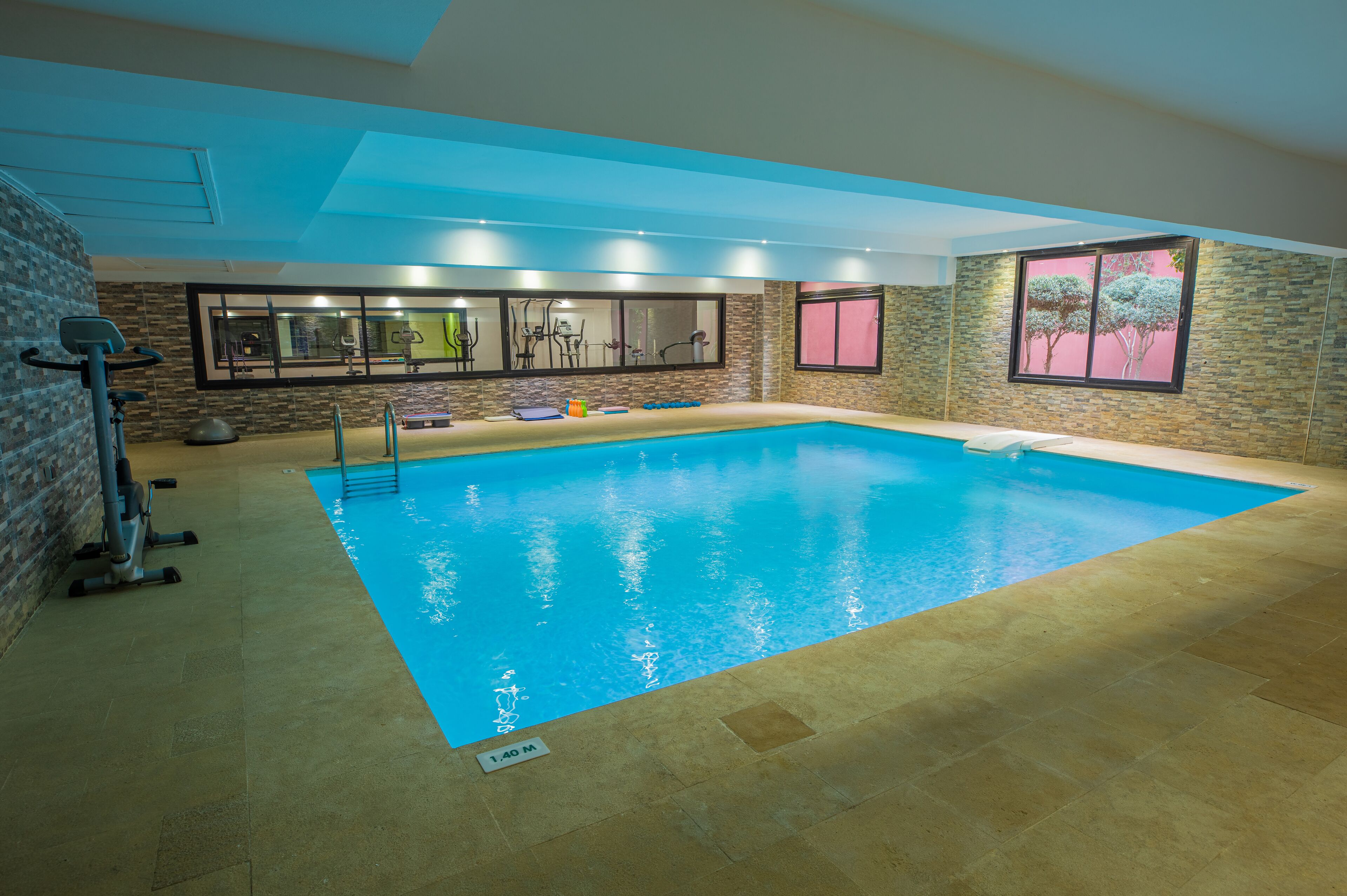 Piscina interior con equipo de ejercicio, una pared de piedra y un ventanal con vistas a un árbol.