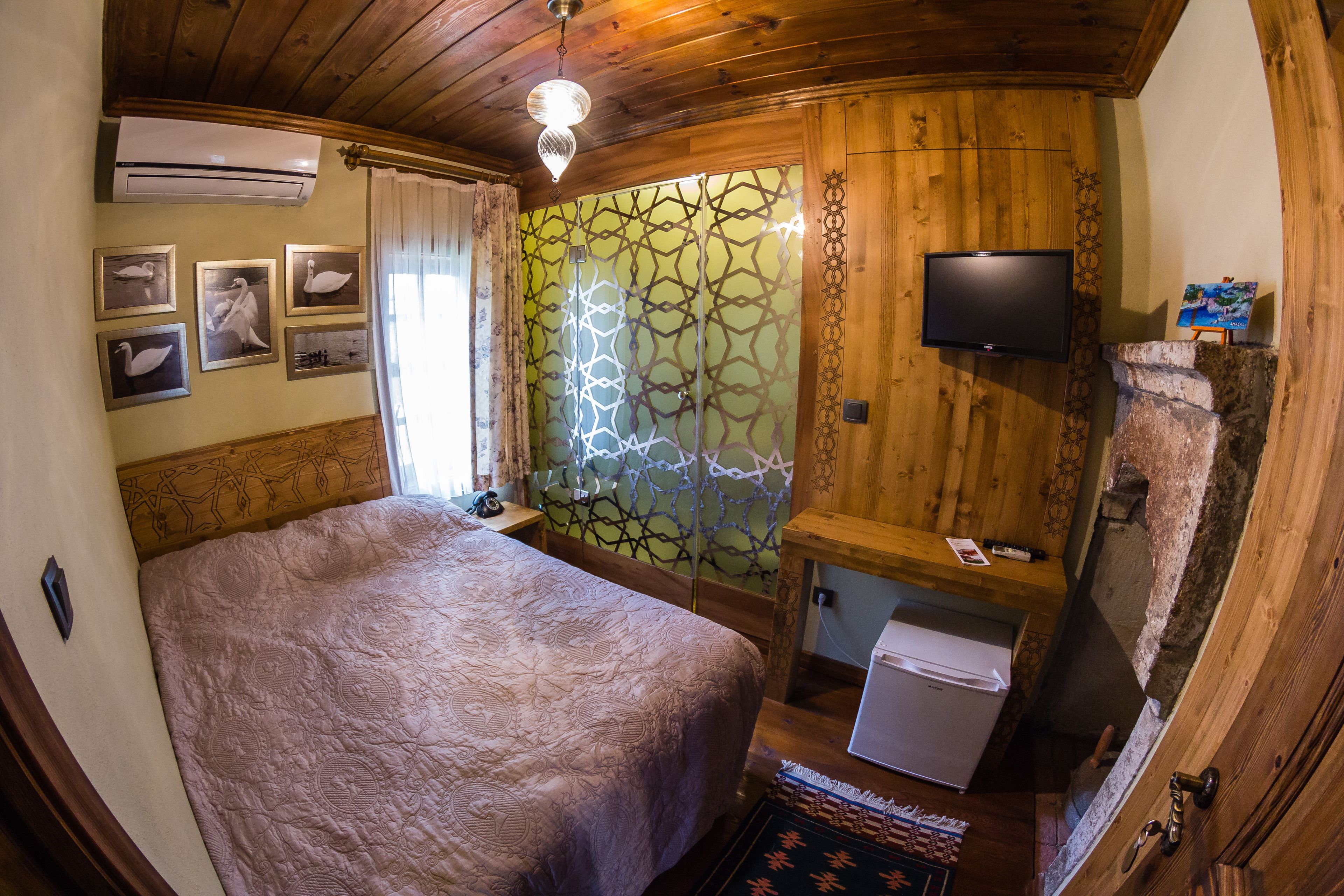 Una habitación con una cama grande, televisión, refrigerador pequeño y una pared de madera con diseño geométrico.