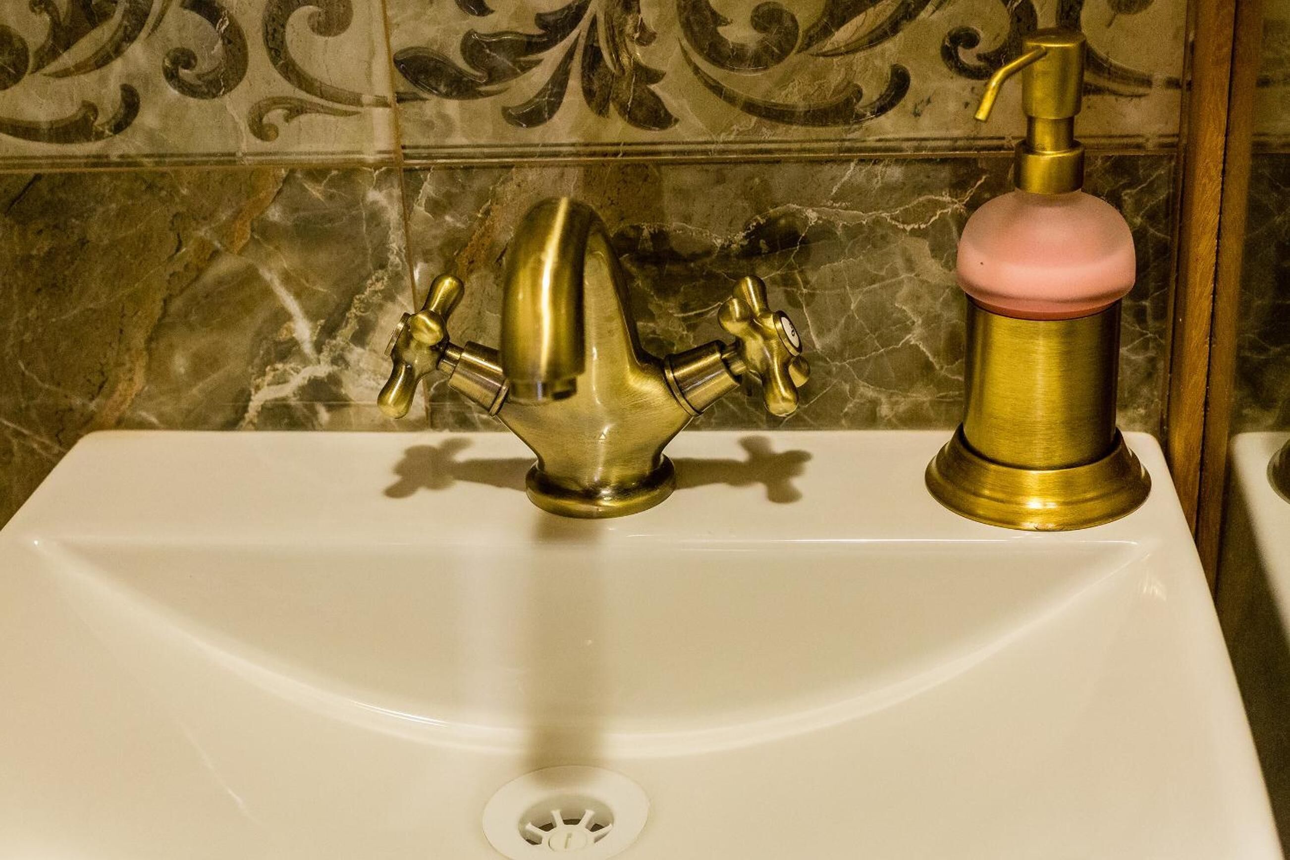 A bathroom sink with two faucets, one brass and one with a pink handle, against a tiled wall.