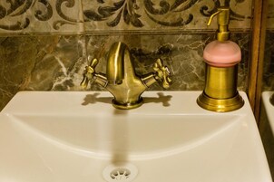 Un lavamanos con dos grifos, uno de bronce y otro con un asa rosa, frente a una pared de azulejos.
