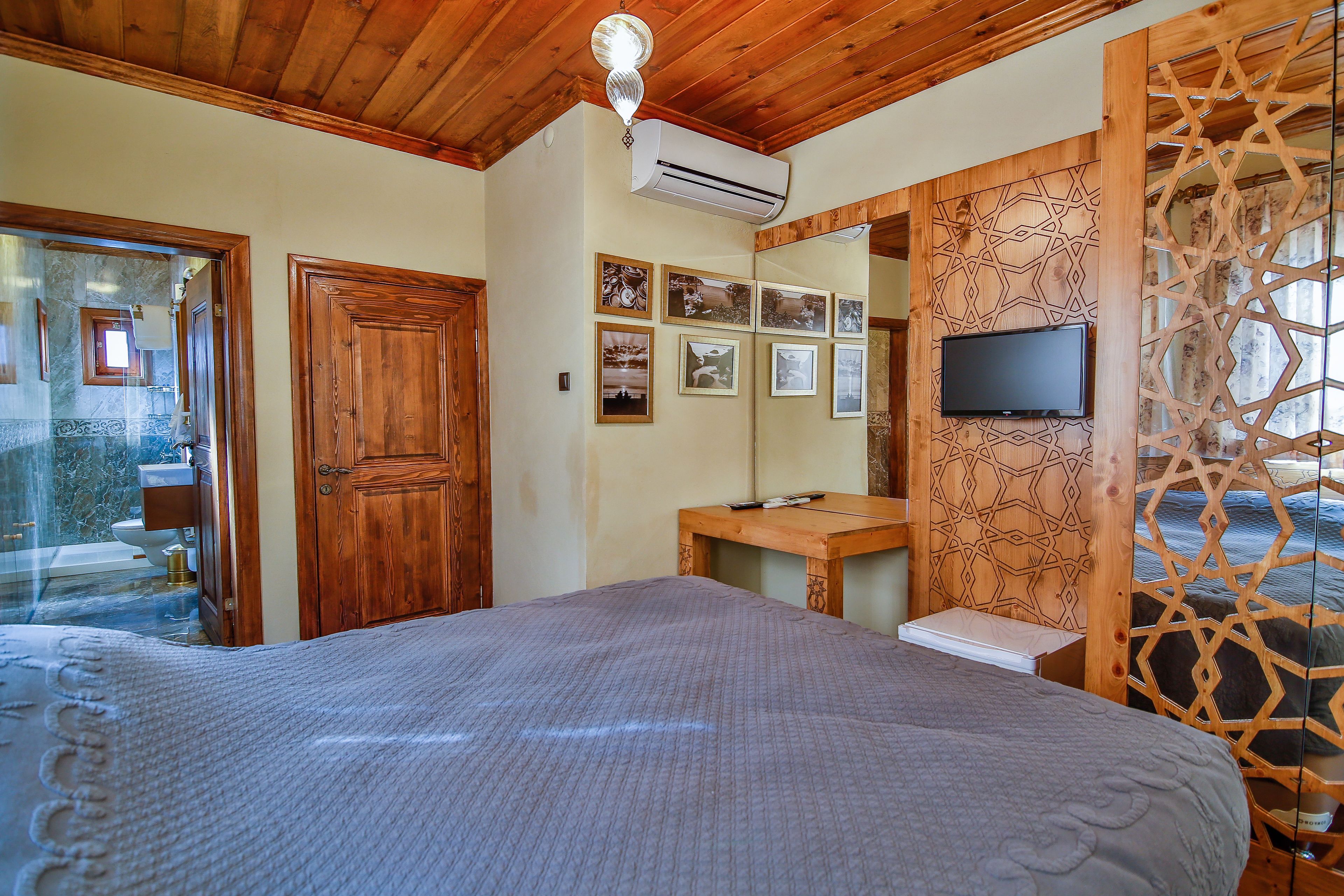A bedroom with a bed, wooden ceiling, air conditioning unit, wooden wall paneling, a television, and a bathroom visible through a glass door.