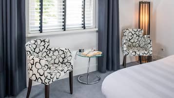A hotel room with a bed, two patterned chairs, a small table with a book and glasses, and a pair of shoes on the floor.