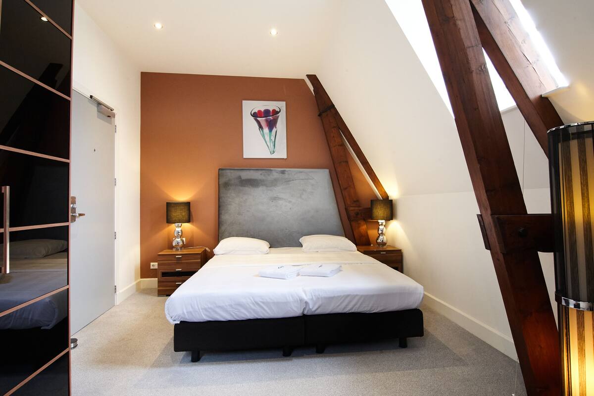 A bedroom with a large bed, two bedside tables with lamps, a headboard, and a painting on the wall.