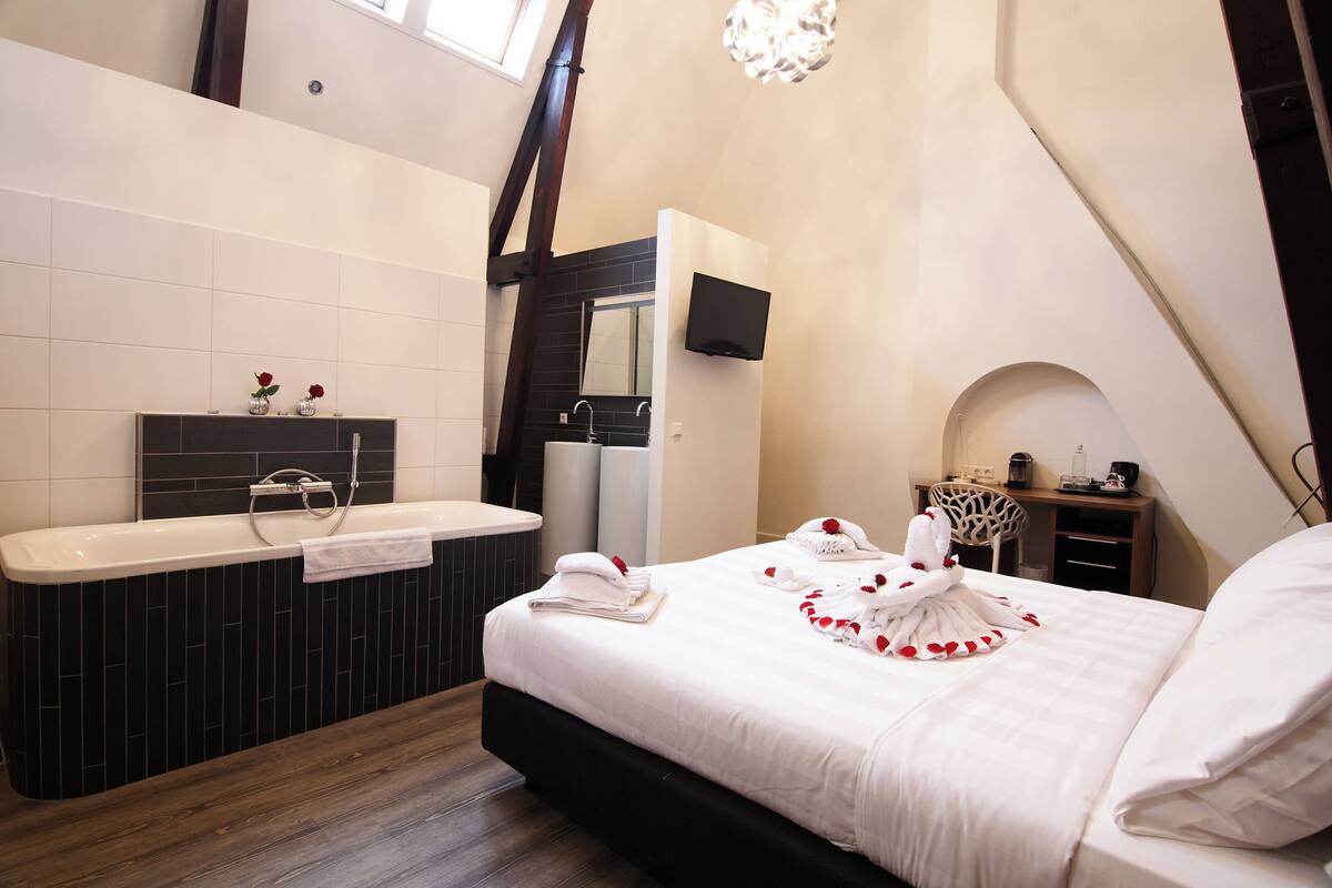 A modern hotel room with a large bed, a bathtub, a flat-screen TV, and a desk with a chair.
