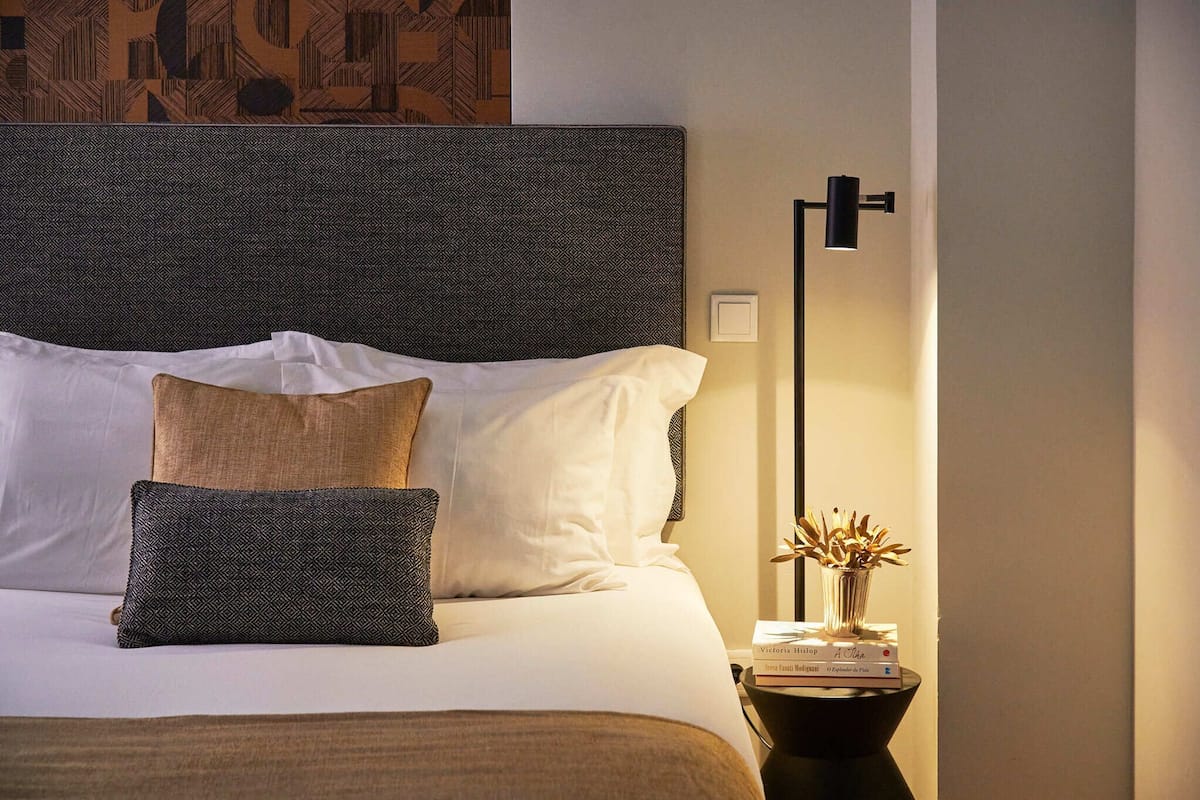 A neatly made bed with white and brown bedding, a patterned headboard, and a bedside table with a lamp and a vase of flowers.