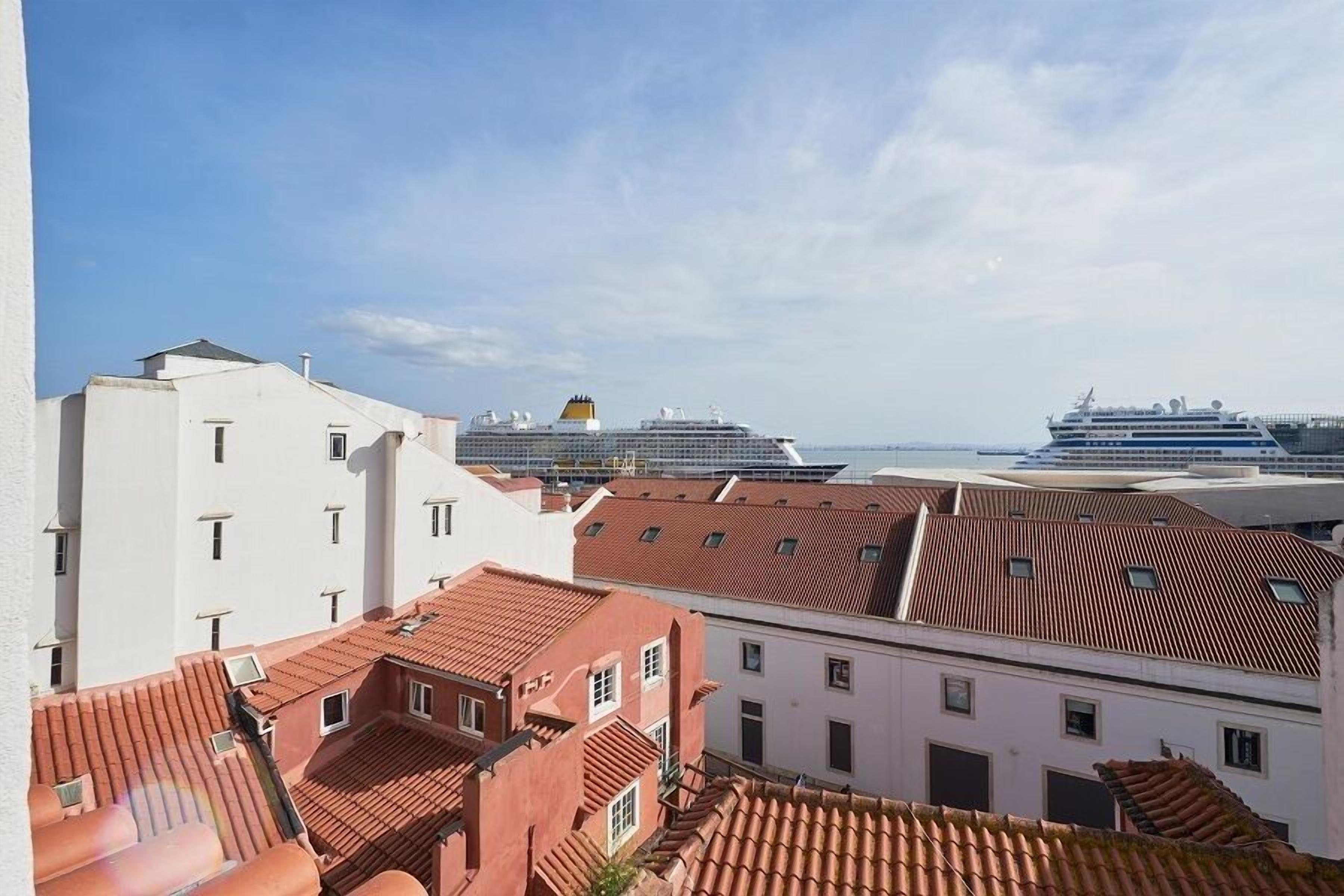 A coastal cityscape with traditional red-tiled roofs and modern buildings, including a large cruise ship docked in the background.