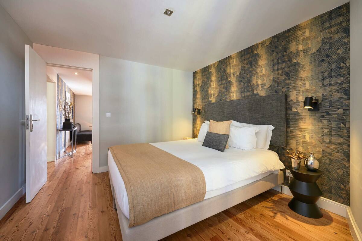 A modern bedroom with a large bed, a headboard, two wall-mounted lights, and a decorative vase on a side table.