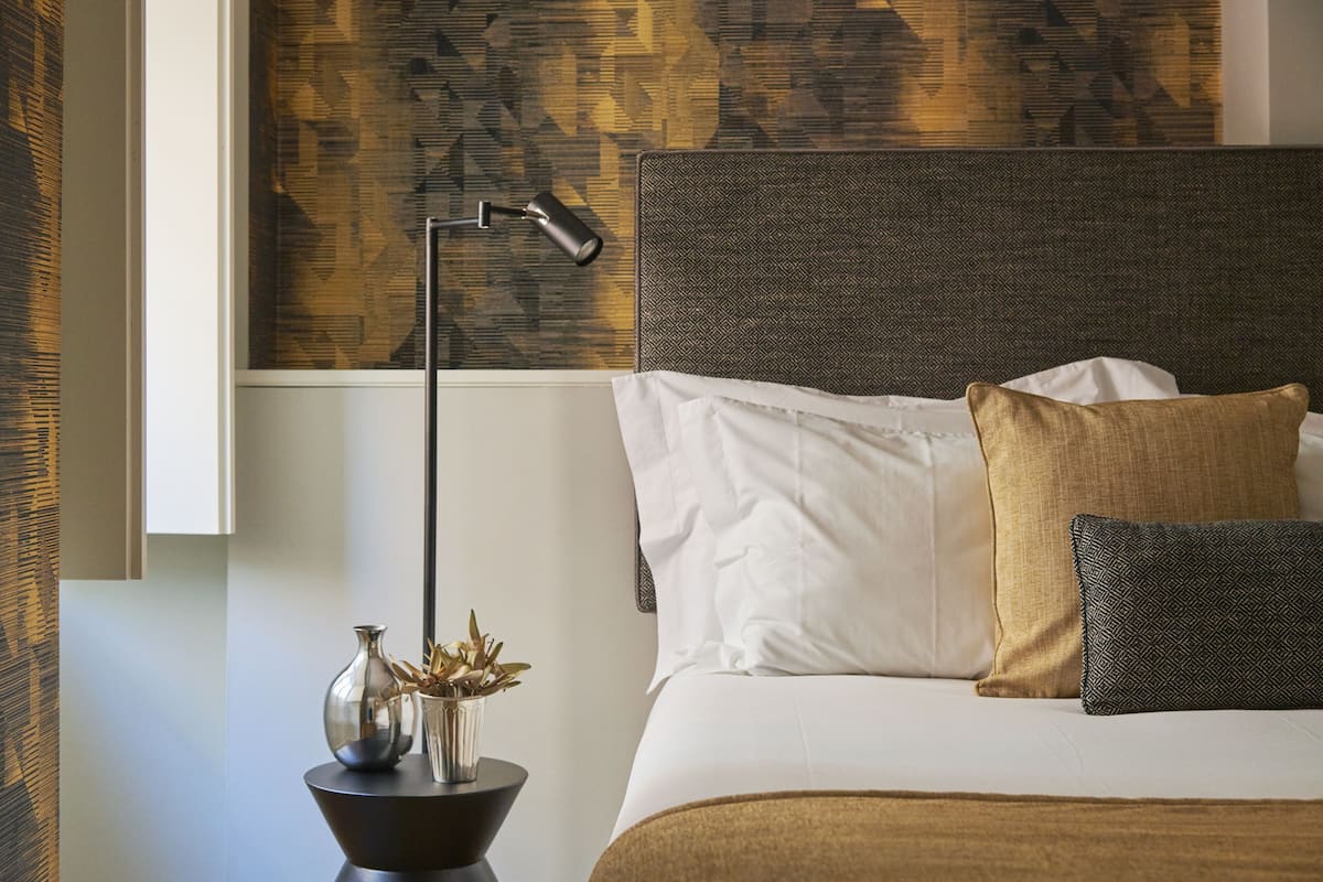 A neatly made bed with white and brown bedding, a modern lamp, and a decorative vase with dried plants on a side table.