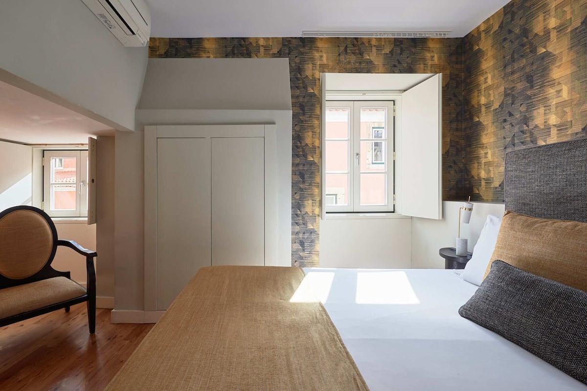 A bedroom with a bed, a chair, a wardrobe, and a window with a view of the outside.
