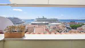 A balcony with a potted plant, overlooking a cityscape with cruise ships and a clear blue sky.
