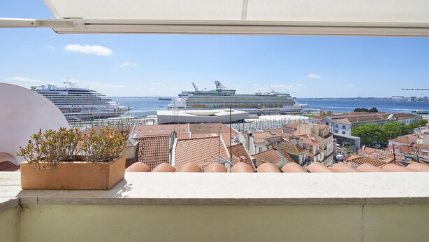 Un balcon agrémenté d’un pot de fleurs, d’où l’on peut admirer un paysage urbain avec des navires de croisière et un ciel bleu dégagé.