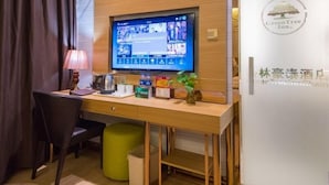 A hotel room with a wooden desk, a television mounted on the wall, a green ottoman, and a trash bin. - GreenTree Inn Nantong Chongchuan District Middle Changjiang Road Express Hotel (Nantong)