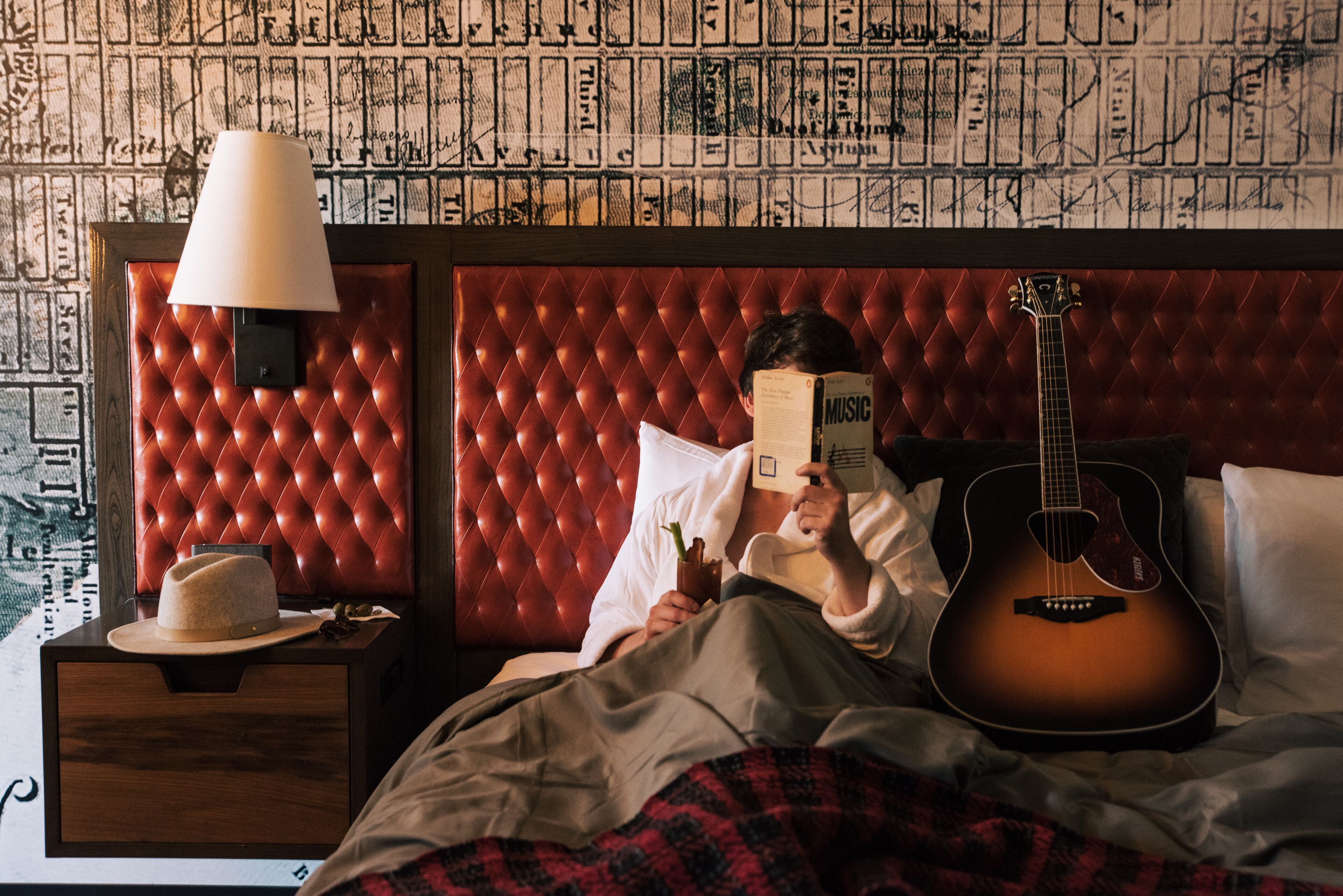 A room with a red tufted headboard, a bedside lamp, a guitar, and a person reading a book.