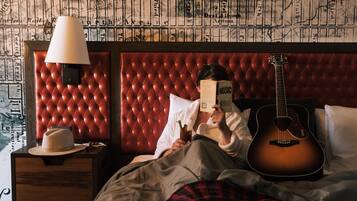 A room with a red tufted headboard, a bedside lamp, a guitar, and a person reading a book.