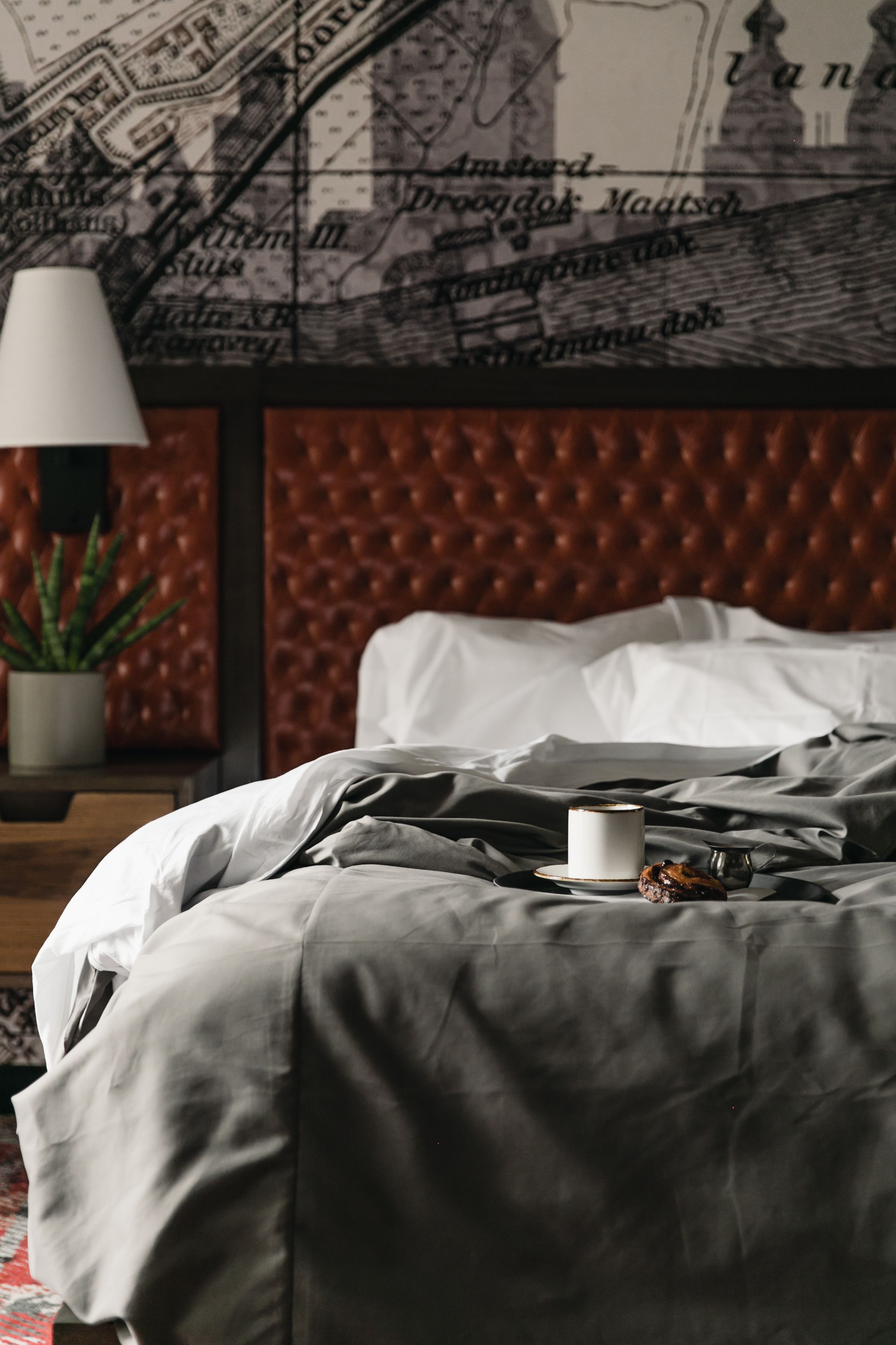 A bedroom with a tufted headboard, a map on the wall, a bedside table with a lamp, and a plant.