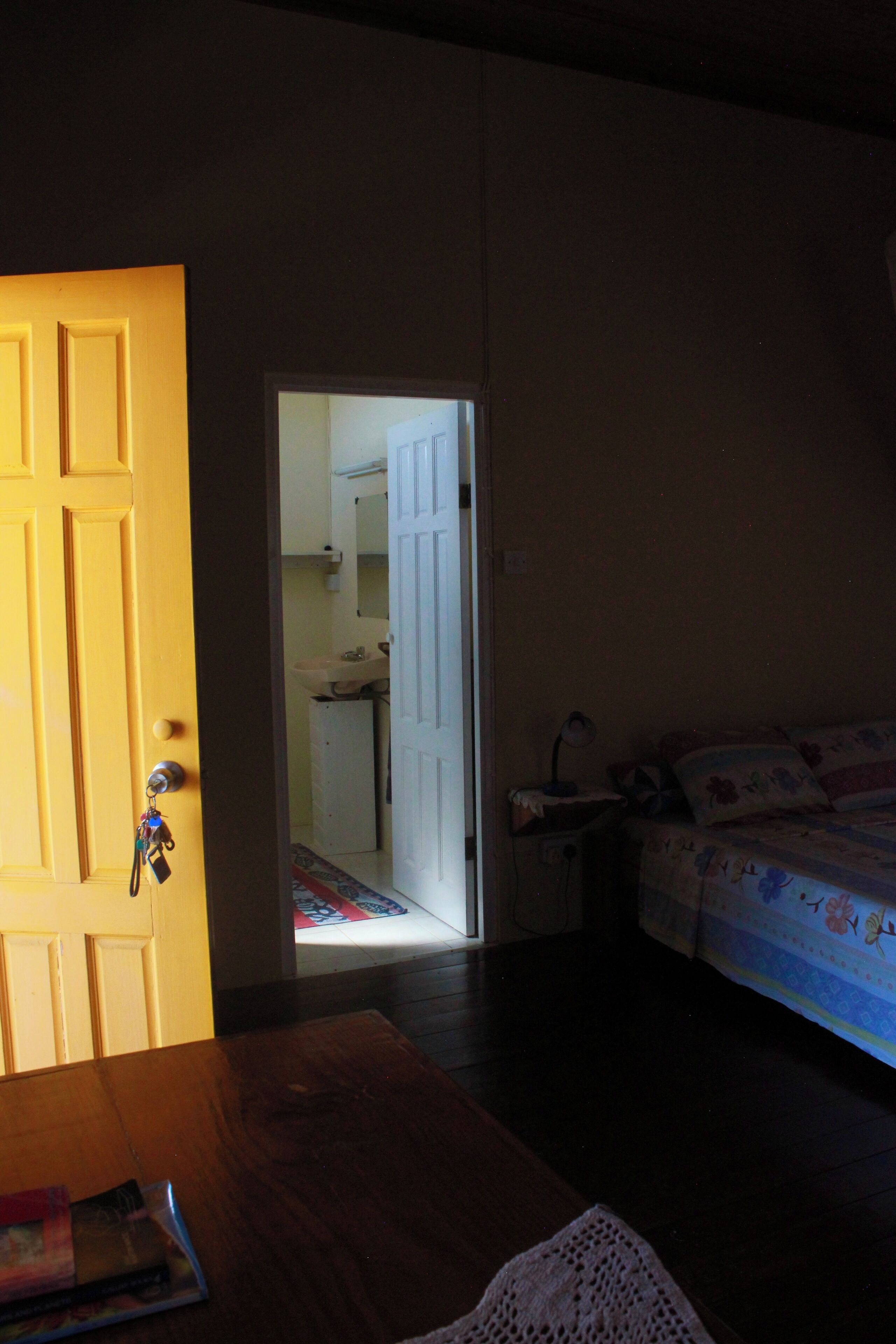 Een kamer met een houten tafel, een bed met bloemenbeddengoed en een deuropening naar een badkamer met een wastafel en spiegel.