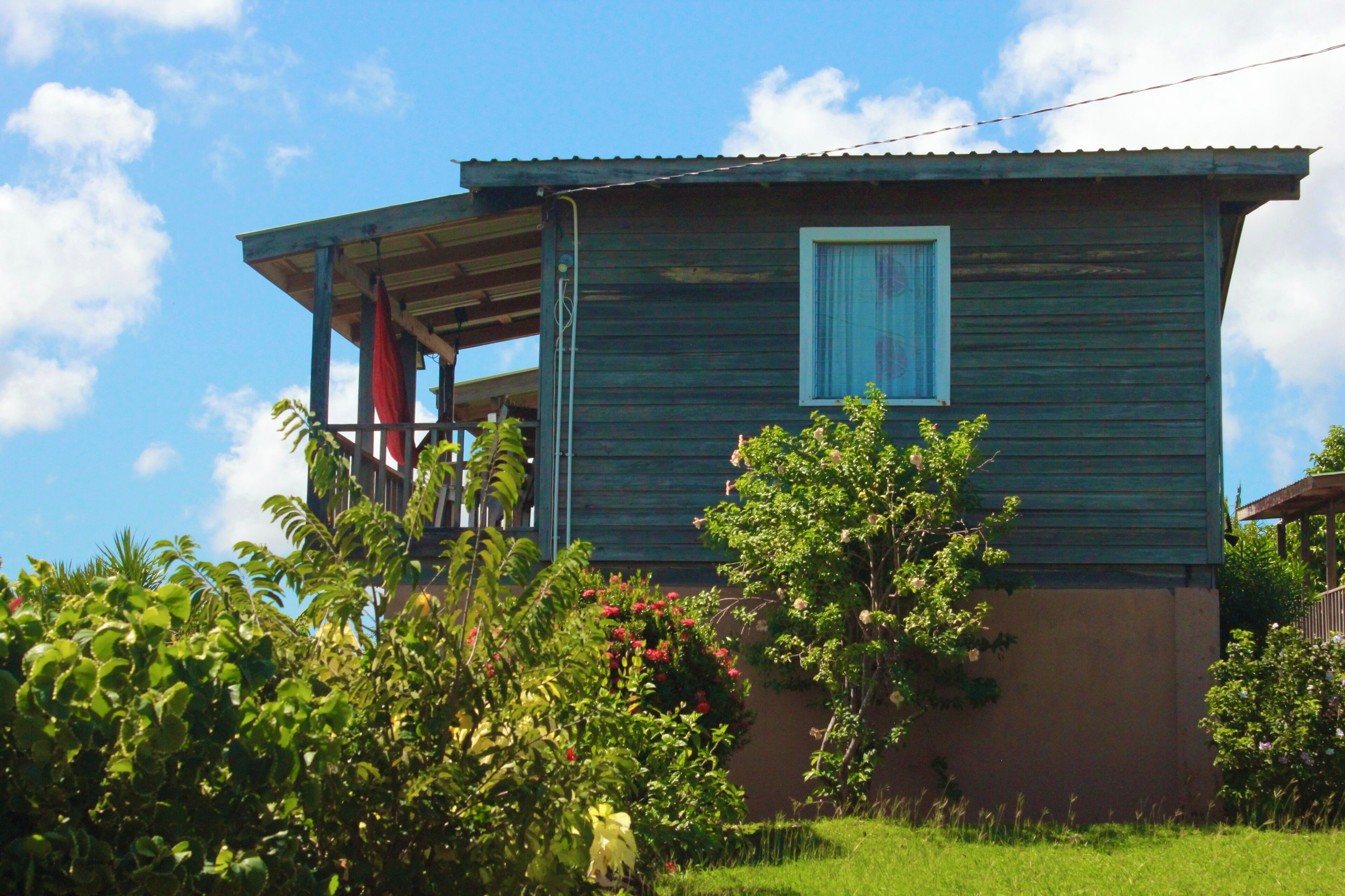 Une maison en bois d’un seul étage, dotée d’un porche et entourée d’une végétation luxuriante et de plantes en fleurs.