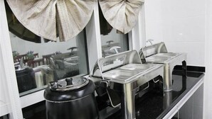 Disposição de buffet com rechauds de aço inoxidável e um bule preto, com uma janela e cortinas ao fundo.