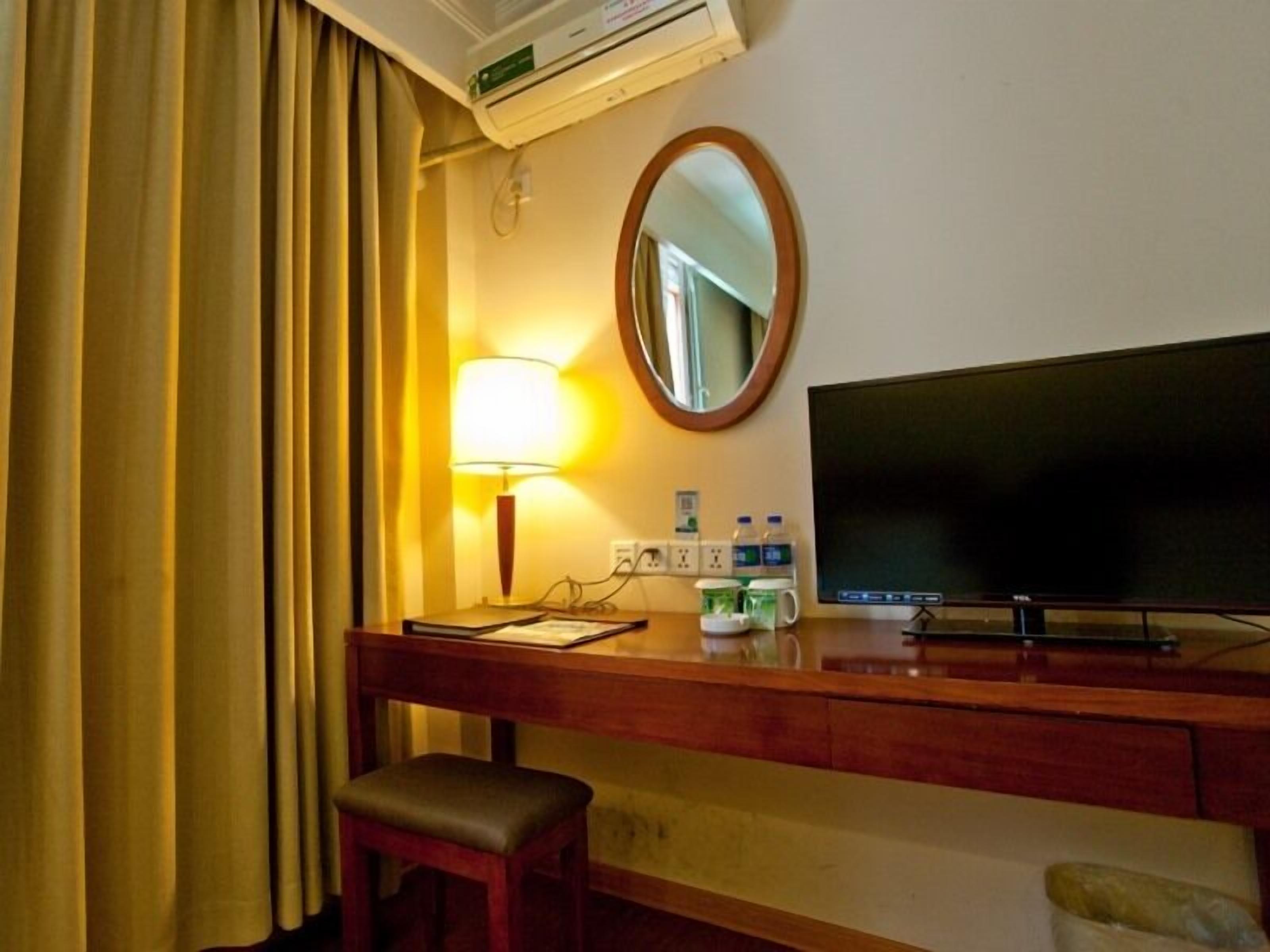 A hotel room with a desk, a television, a mirror, a lamp, and a window with curtains.