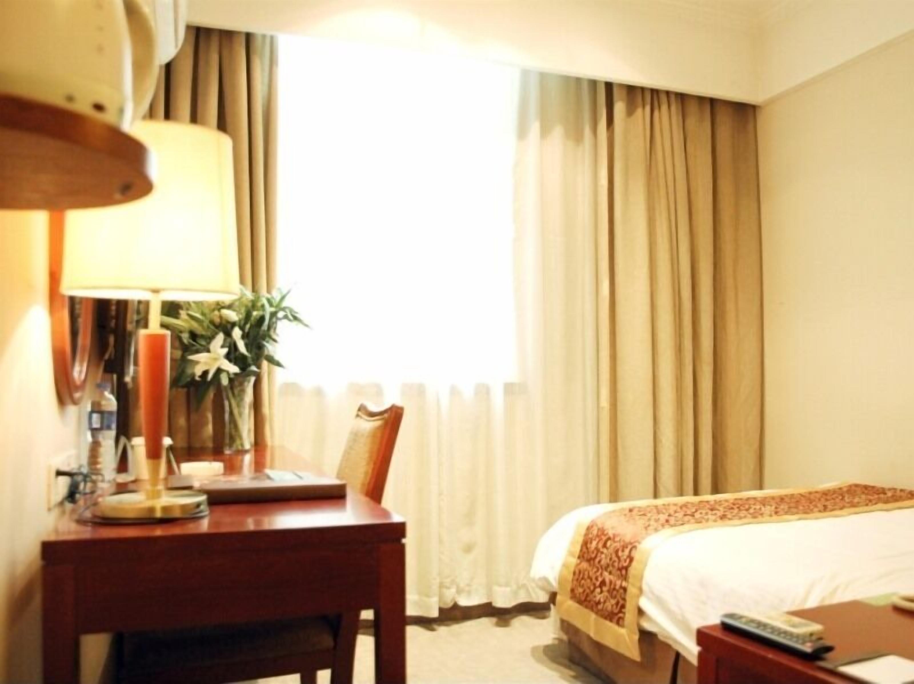 Habitación de hotel con cama, escritorio con lámpara, florero con flores y ventana con cortinas.