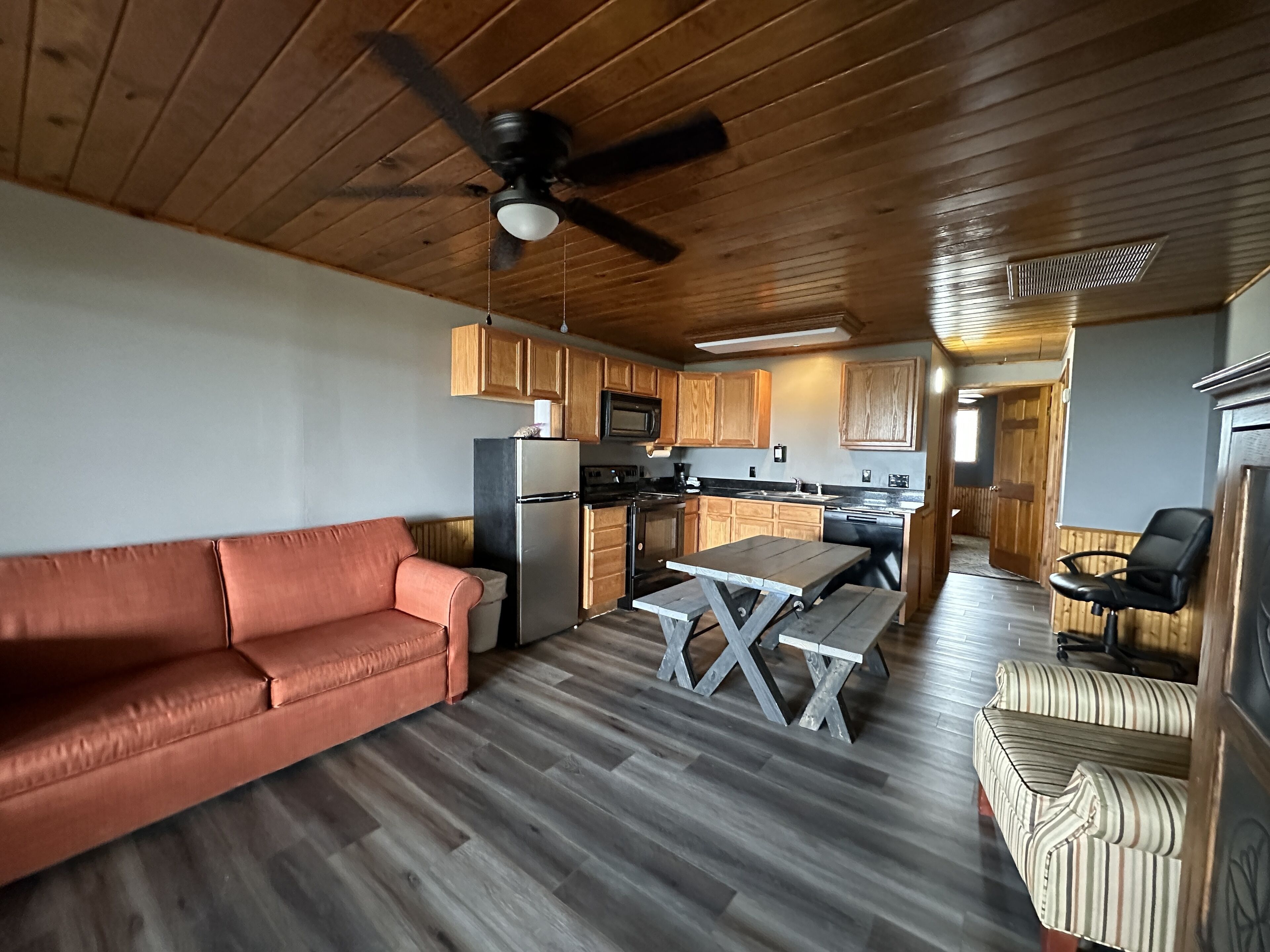 A spacious living area with a wooden ceiling, a ceiling fan, a sofa, a dining table, and a kitchen with wooden cabinets and appliances.
