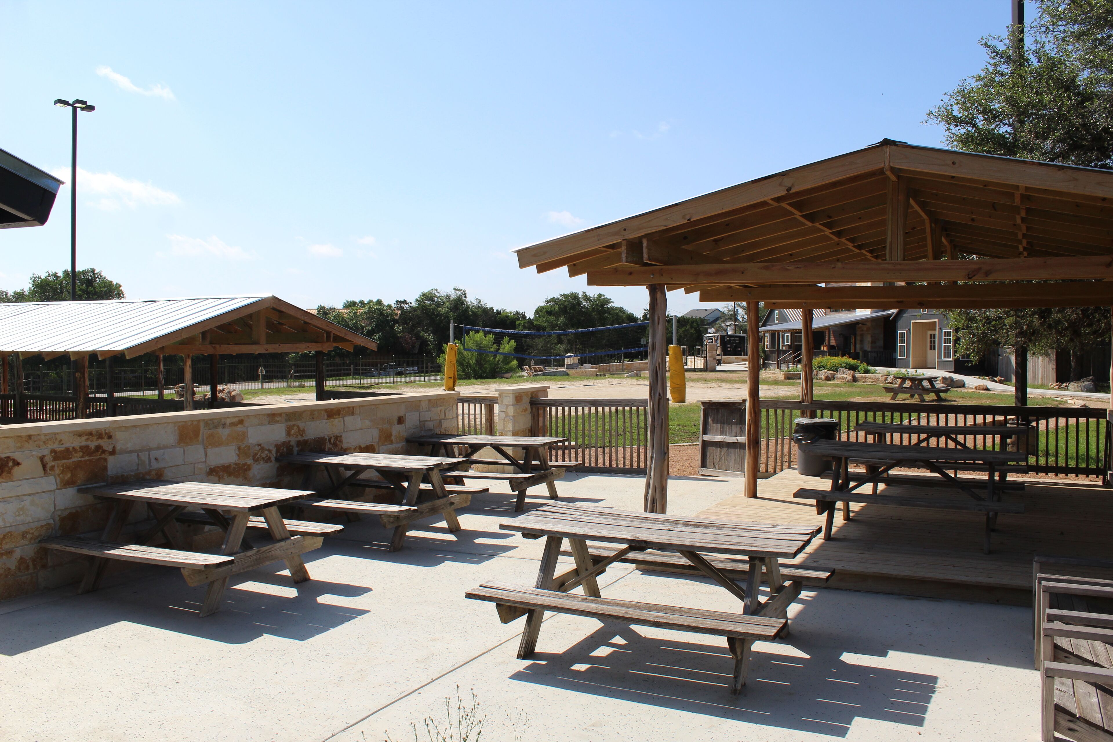 A covered picnic area with multiple wooden picnic tables and benches, a stone wall, and a metal roof structure.