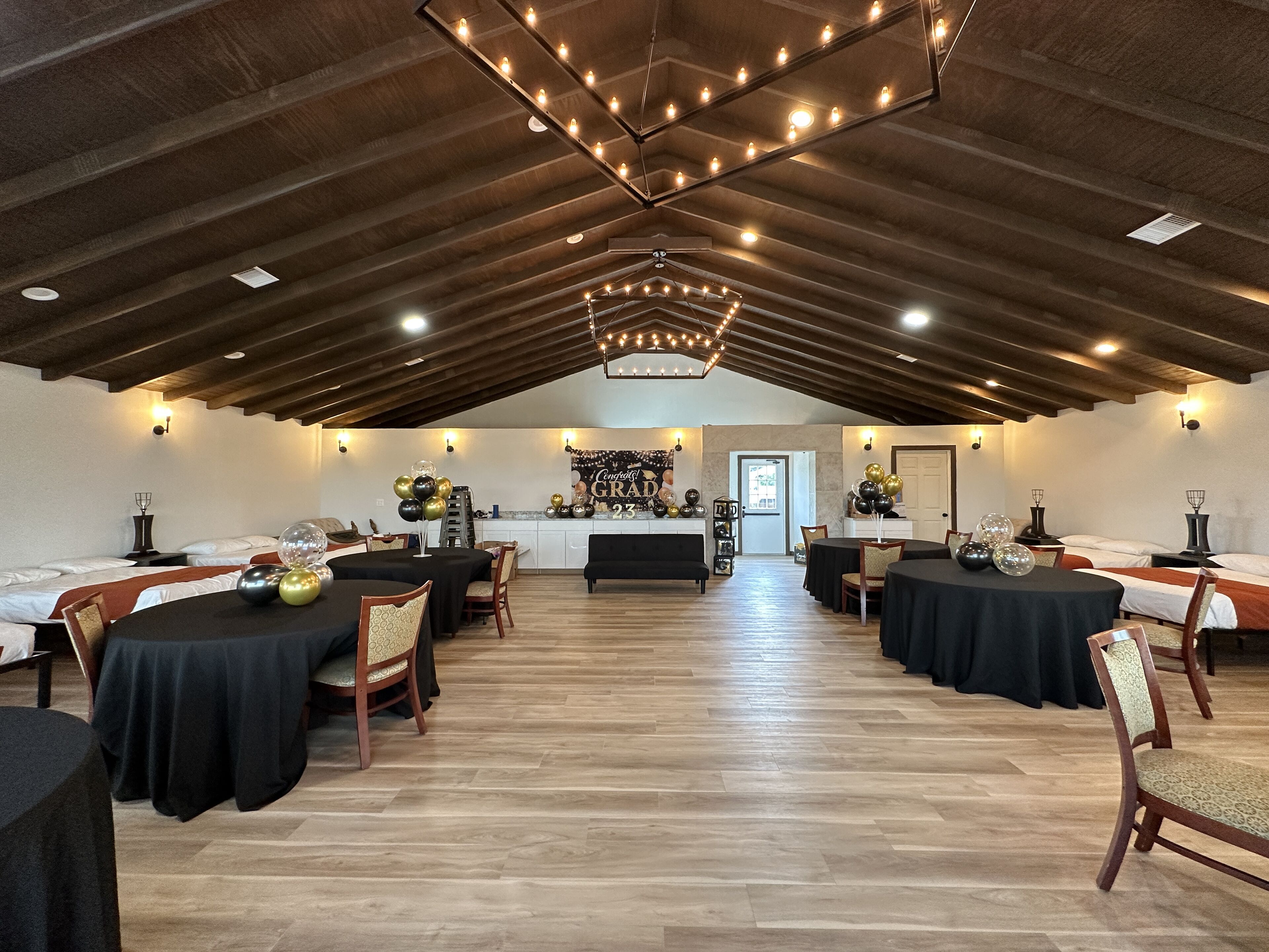 Une pièce spacieuse avec un plafond haut, des poutres en bois et un lustre central. Des tables sont disposées dans la pièce, et des chaises avec des coussins sont également présentes.