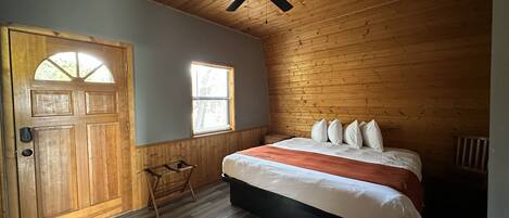 Ein Schlafzimmer mit Holzbalkendecke, einem Bett mit weißer und orangefarbener Bettwäsche, einem Deckenventilator und einem Fenster mit Jalousien.