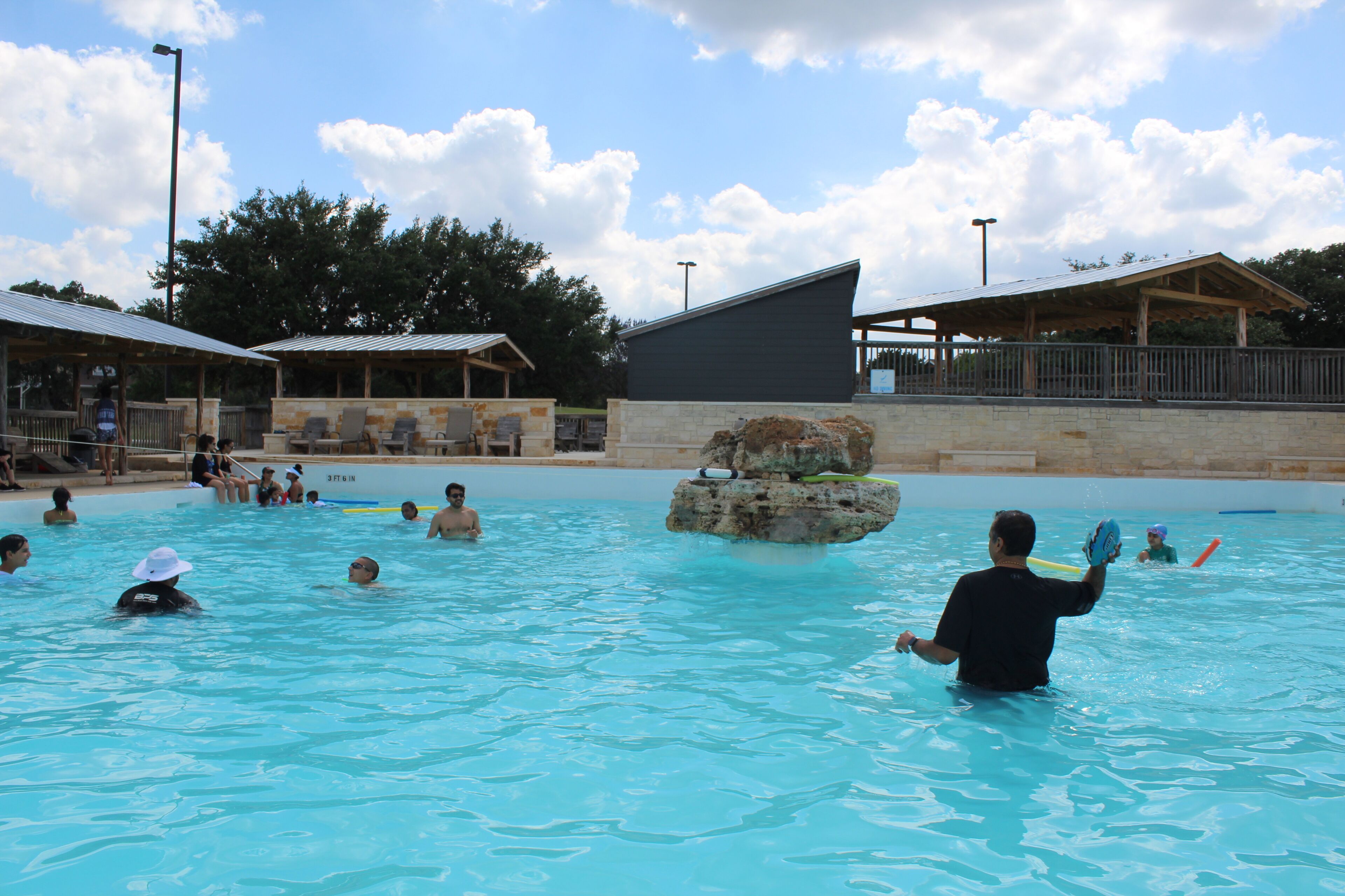 A swimming pool with people enjoying the water, surrounded by buildings and trees.