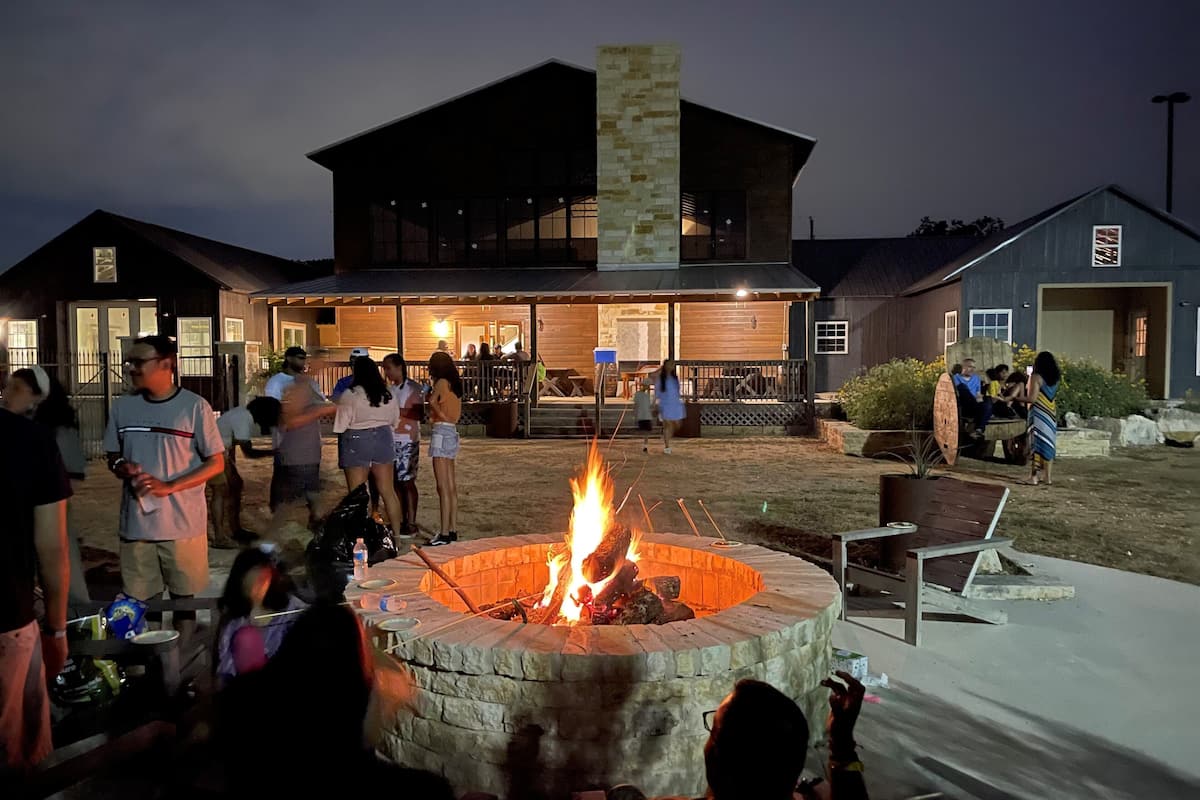 A group of people gathered around a fire pit in an outdoor setting with a building in the background.