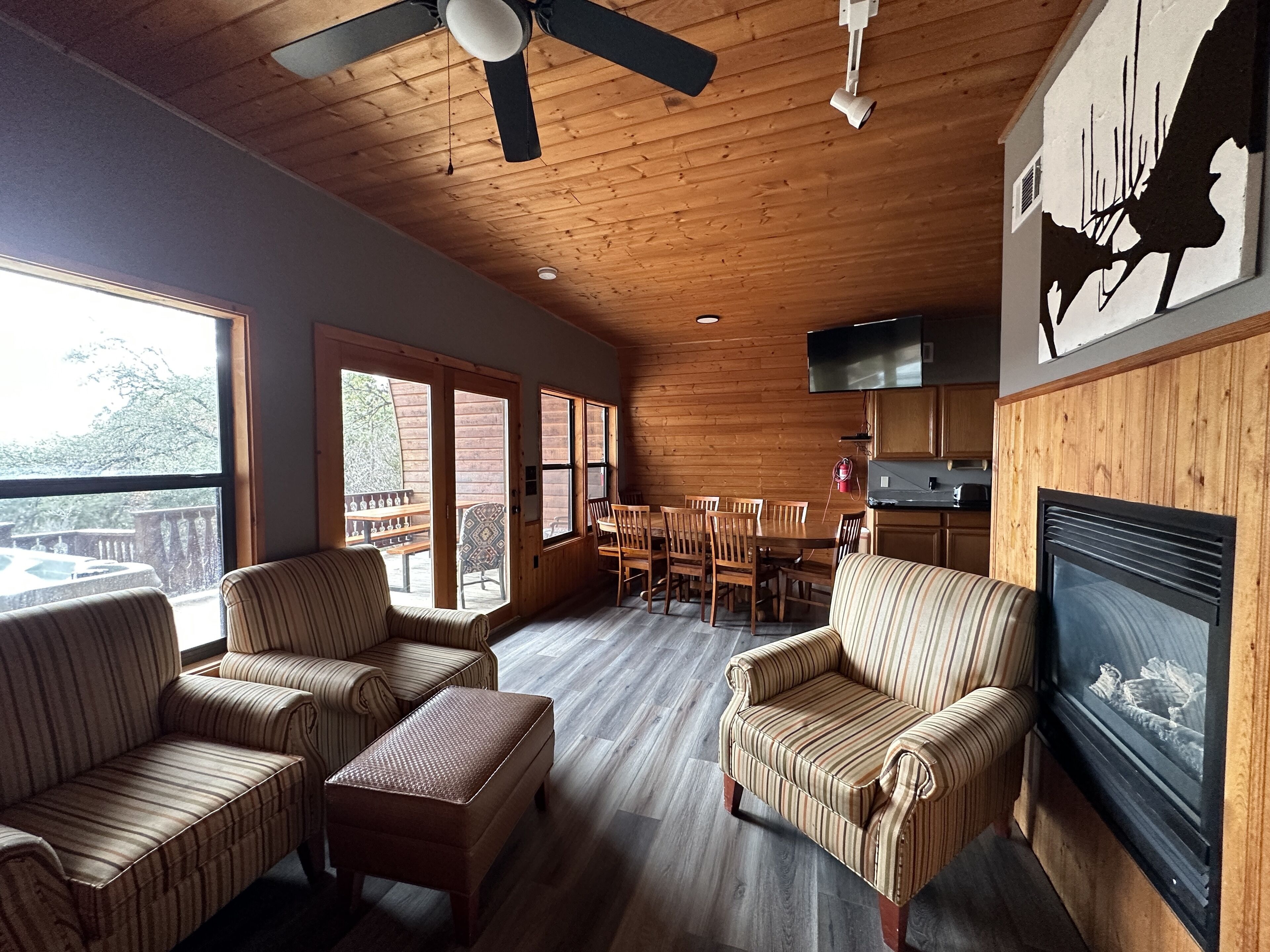 A spacious living room with wooden floors, a ceiling fan, and a fireplace. There are two sofas and a dining table with chairs.