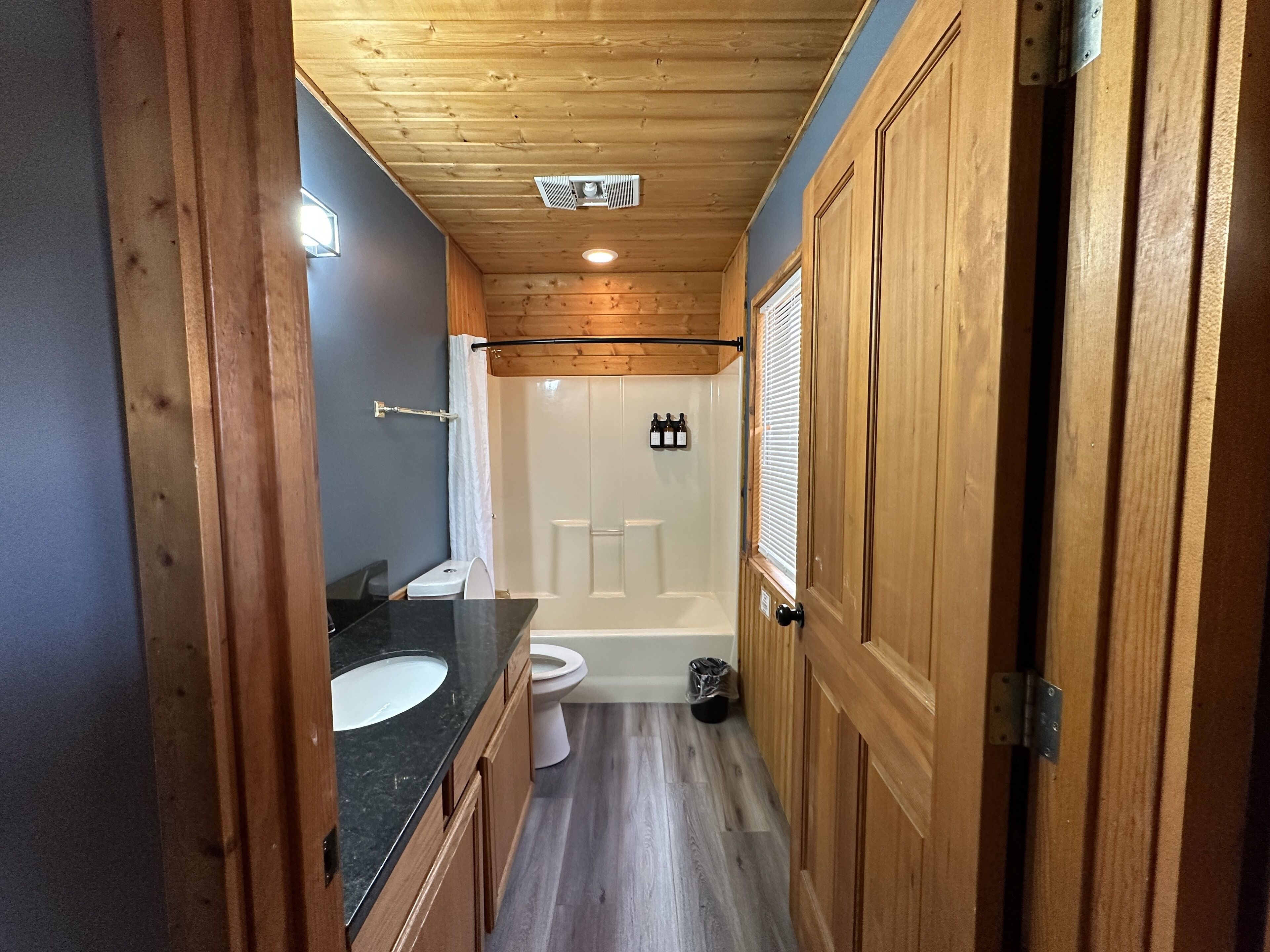 Un baño estrecho con techo de madera, un lavamanos con encimera negra, un inodoro blanco y una zona de ducha.