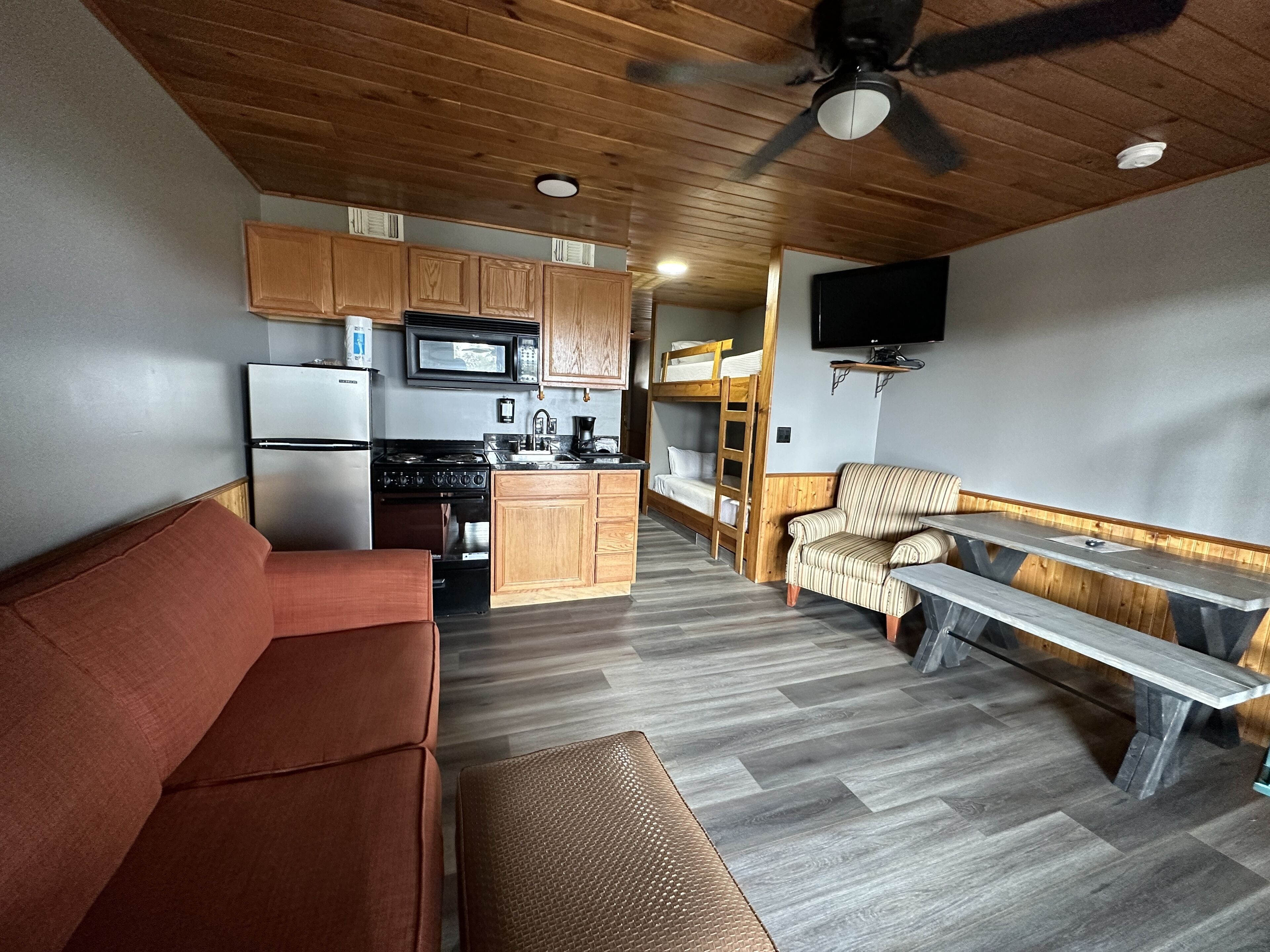 A compact kitchen with wooden cabinets, a microwave, and a stove. There is a sofa and a bench in the living area, and a TV mounted on the wall.