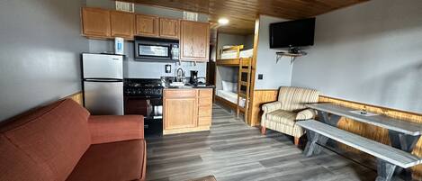 A compact kitchen with wooden cabinets, a microwave, and a stove. There is a sofa and a bench in the living area, and a TV mounted on the wall.