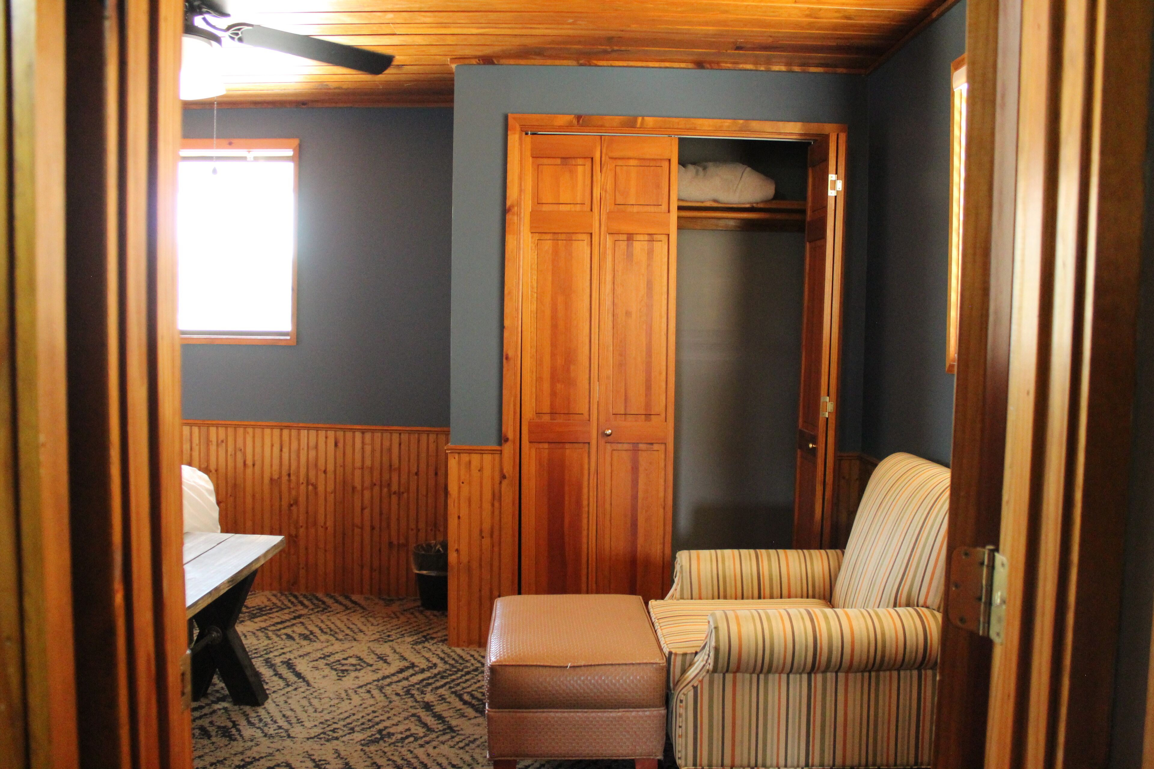 A room with a wooden wardrobe, a striped armchair, a small table, and a window.