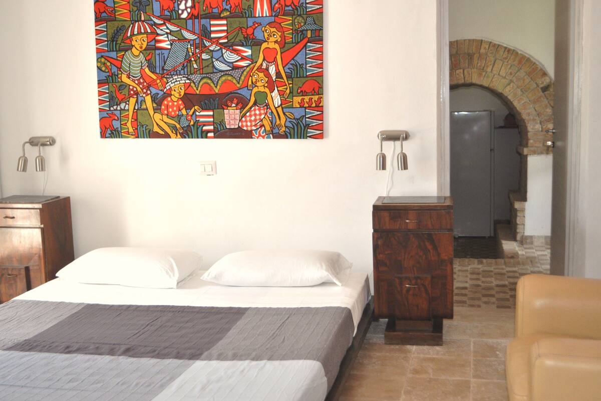 A bedroom with a bed, a nightstand, a dresser, a colorful painting, and a doorway leading to a bathroom.