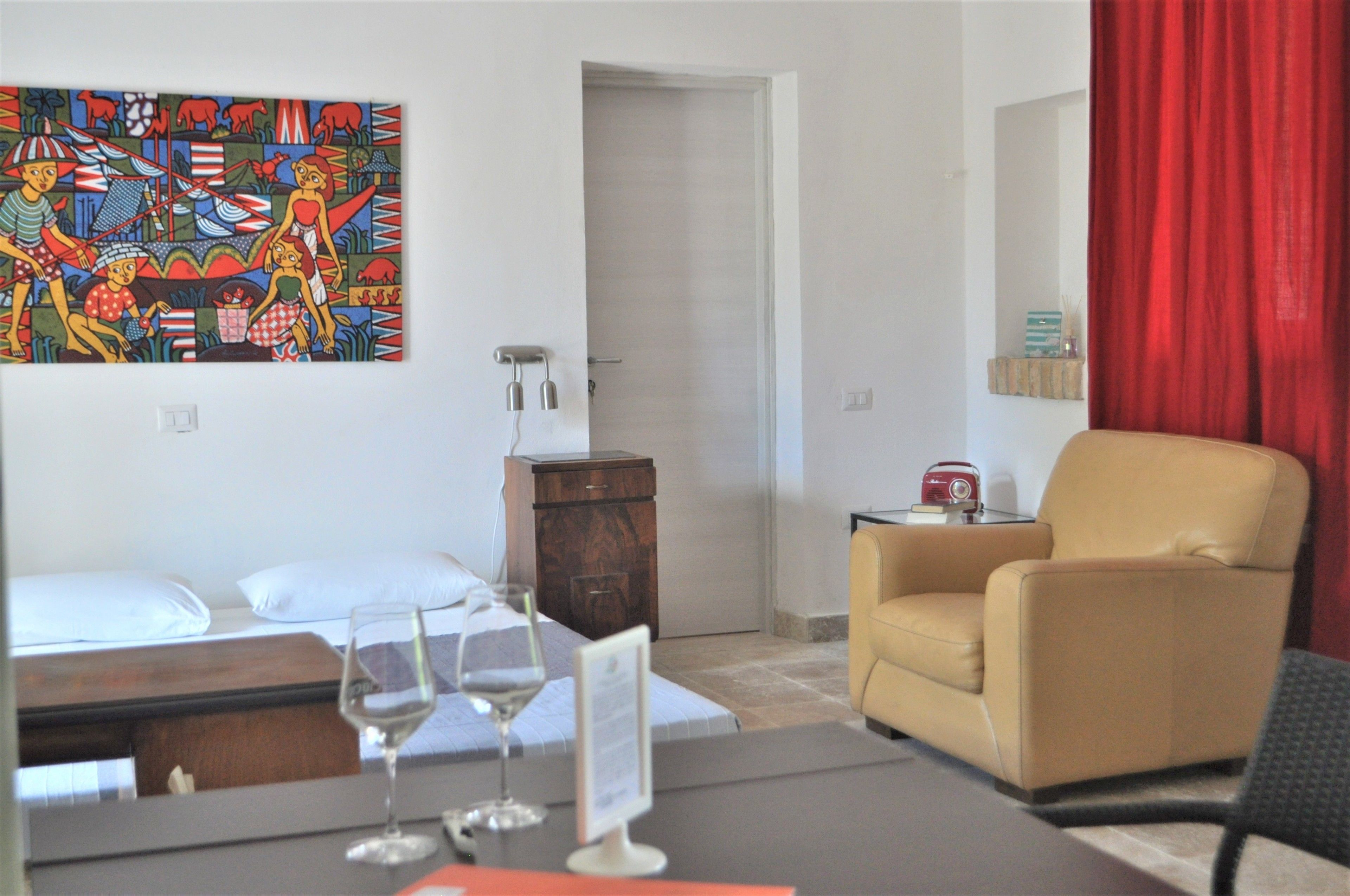 Une chambre d’hôtel avec un lit, un bureau sur lequel se trouve un verre à vin, une chaise, un rideau rouge, un tableau accroché au mur et une petite table avec une radio.
